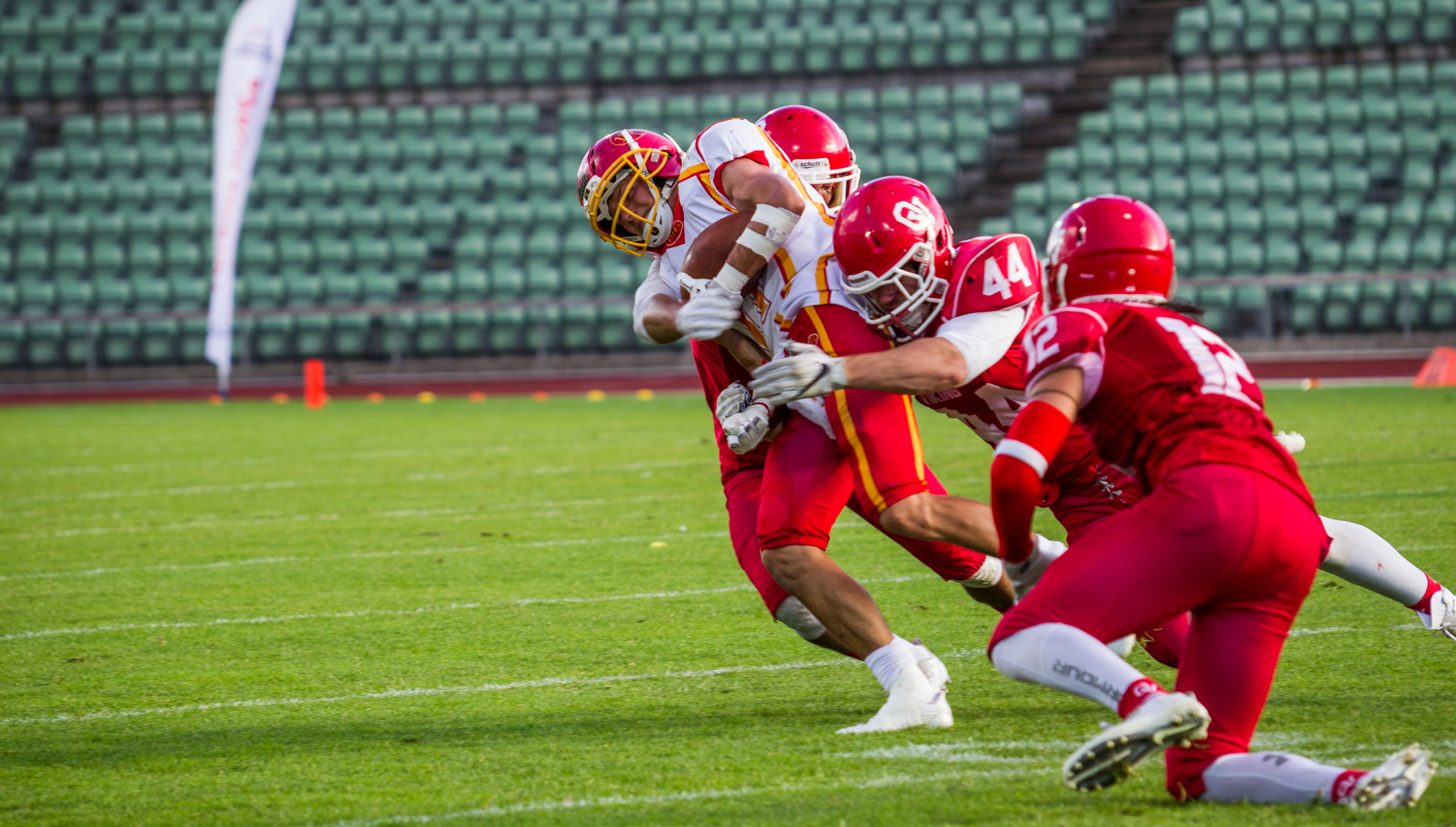 Several american football players on the field. One has the ball and is being tackled by two opponents.