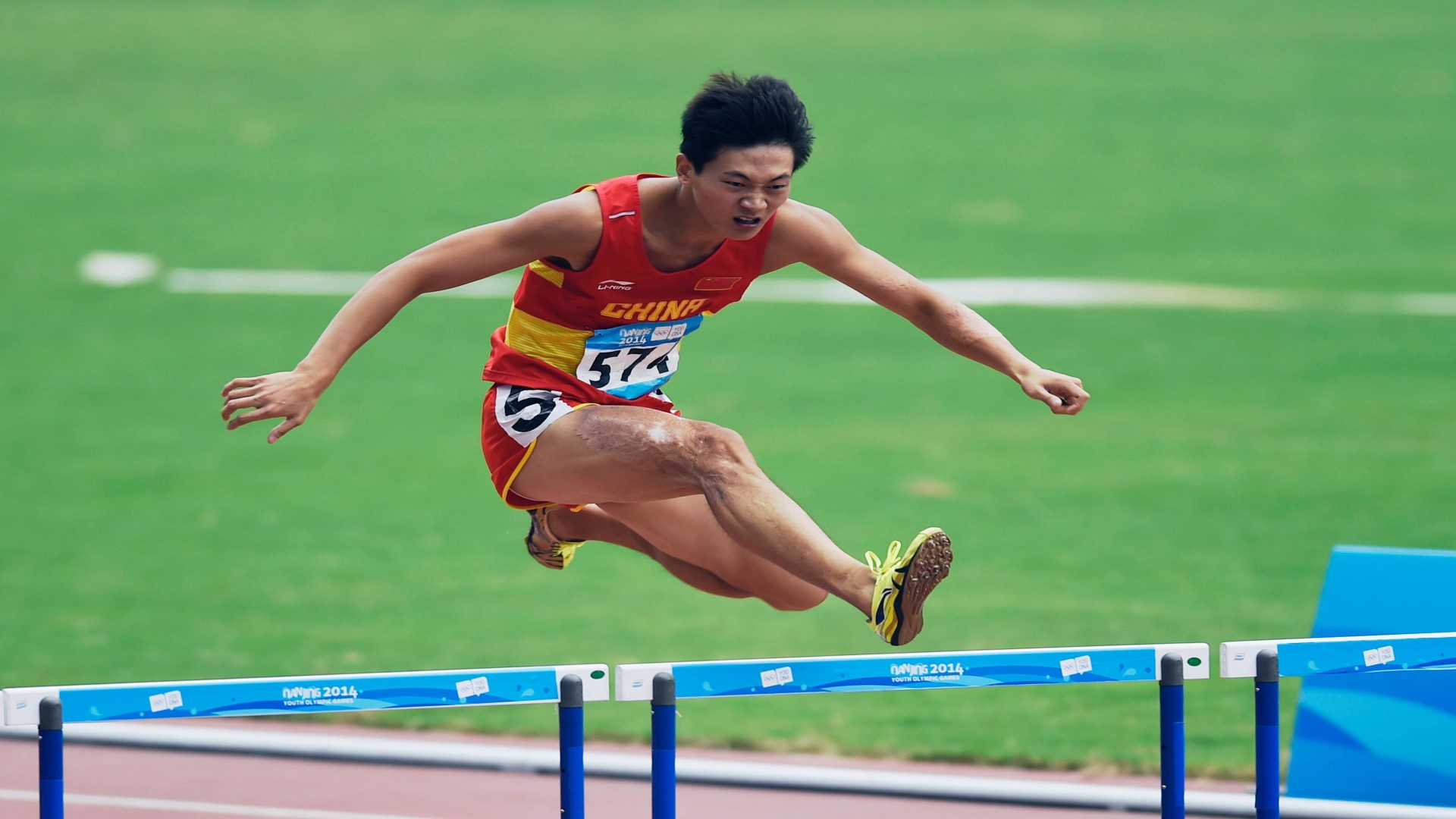 Athlete on the track and field in a hurdle competition. Athlete is in the air, his right leg infront as he is jumping the hurdle.