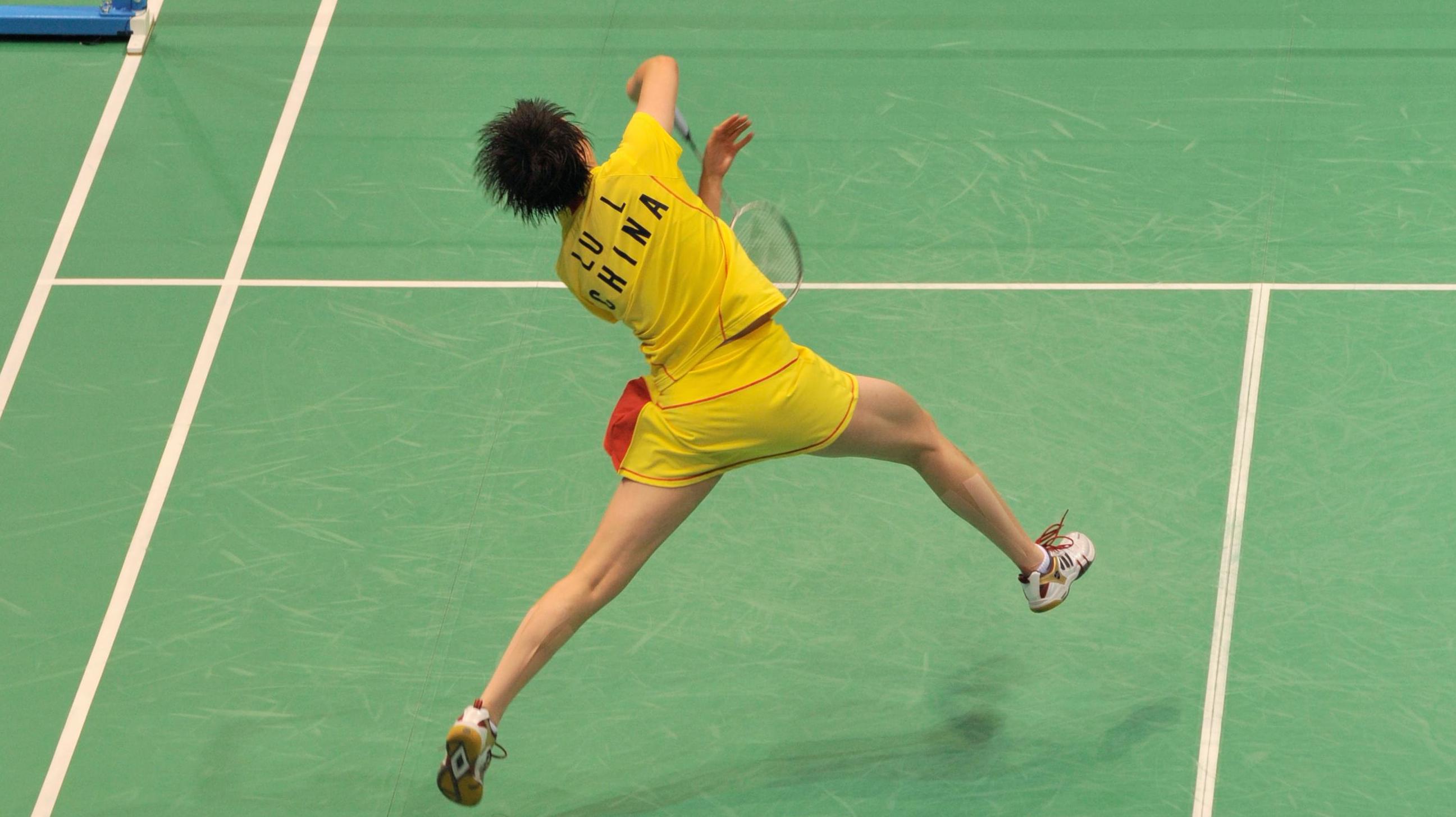 Badminton player one the court seen from behind. Athlete in the air trying to hit with the racket.