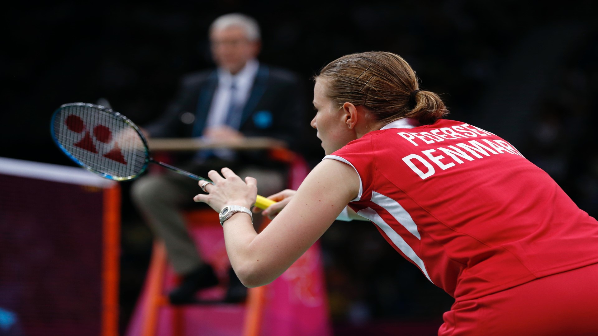 Player seen from the side, slightly bendt forward with racket in her hand, eyes forward.