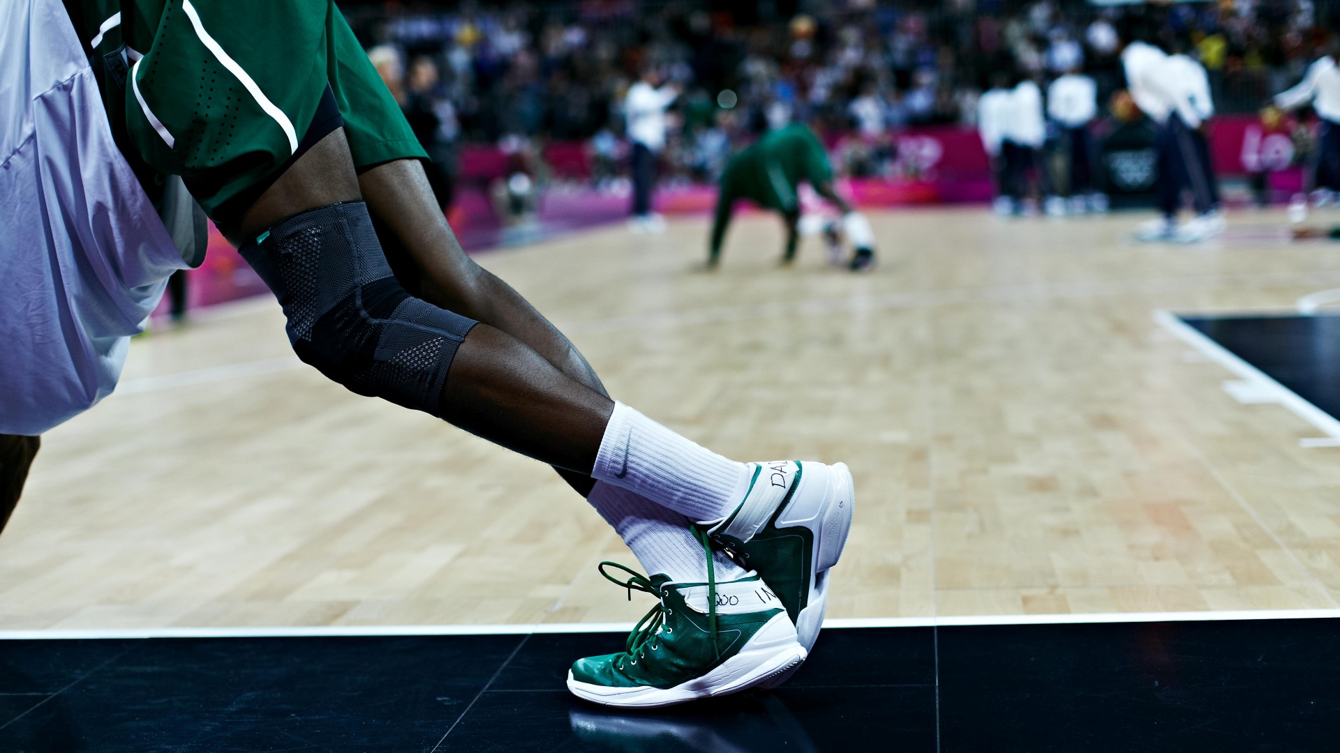Close up of a basketballplayer on the side of the court, stretching, with one foot on the ground and the other foot behind the first one. 