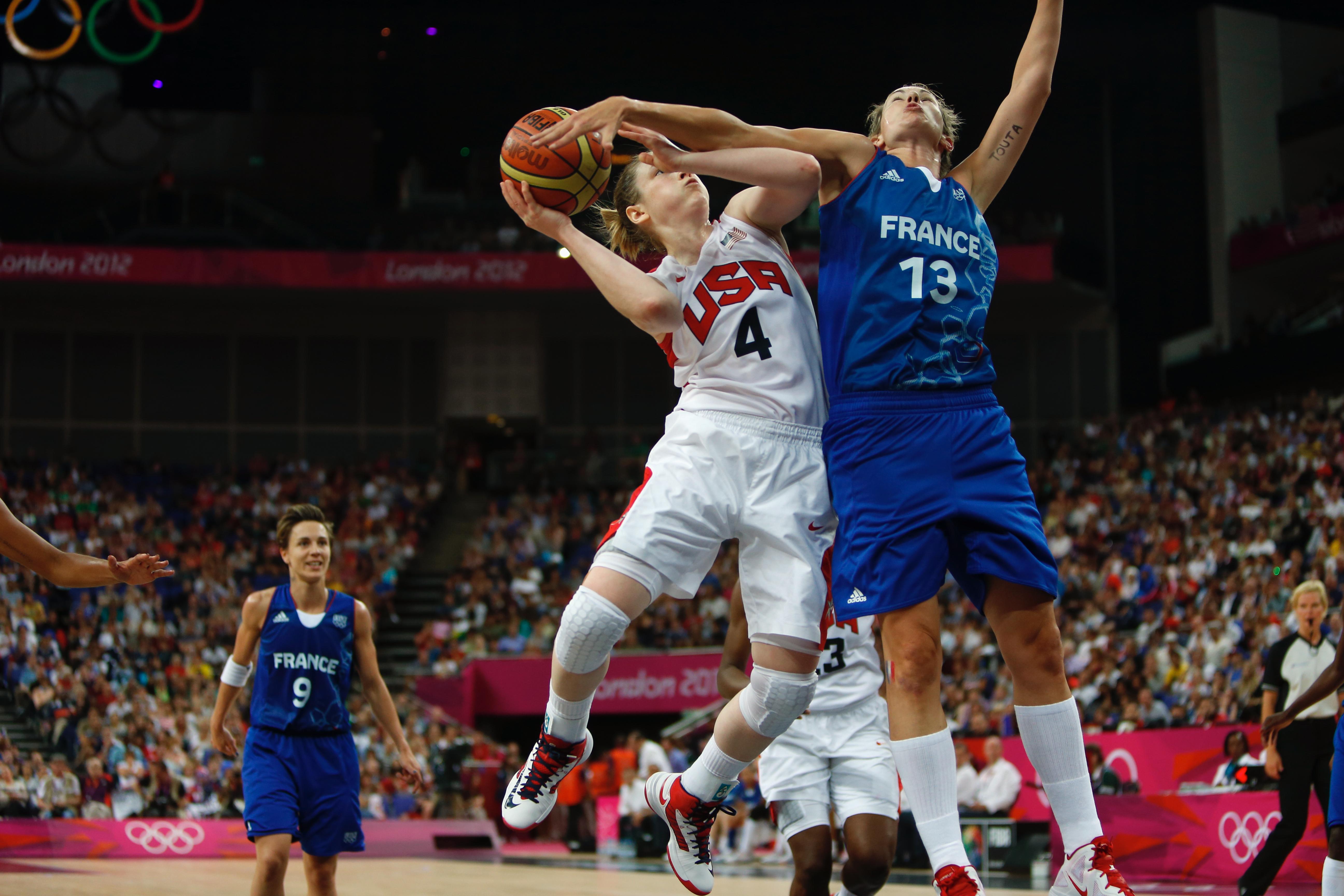 Basketballgame in the Olympics, on the court. Two athletes in the air, one athlete has the ball in her right hand, the other players is trying to block.