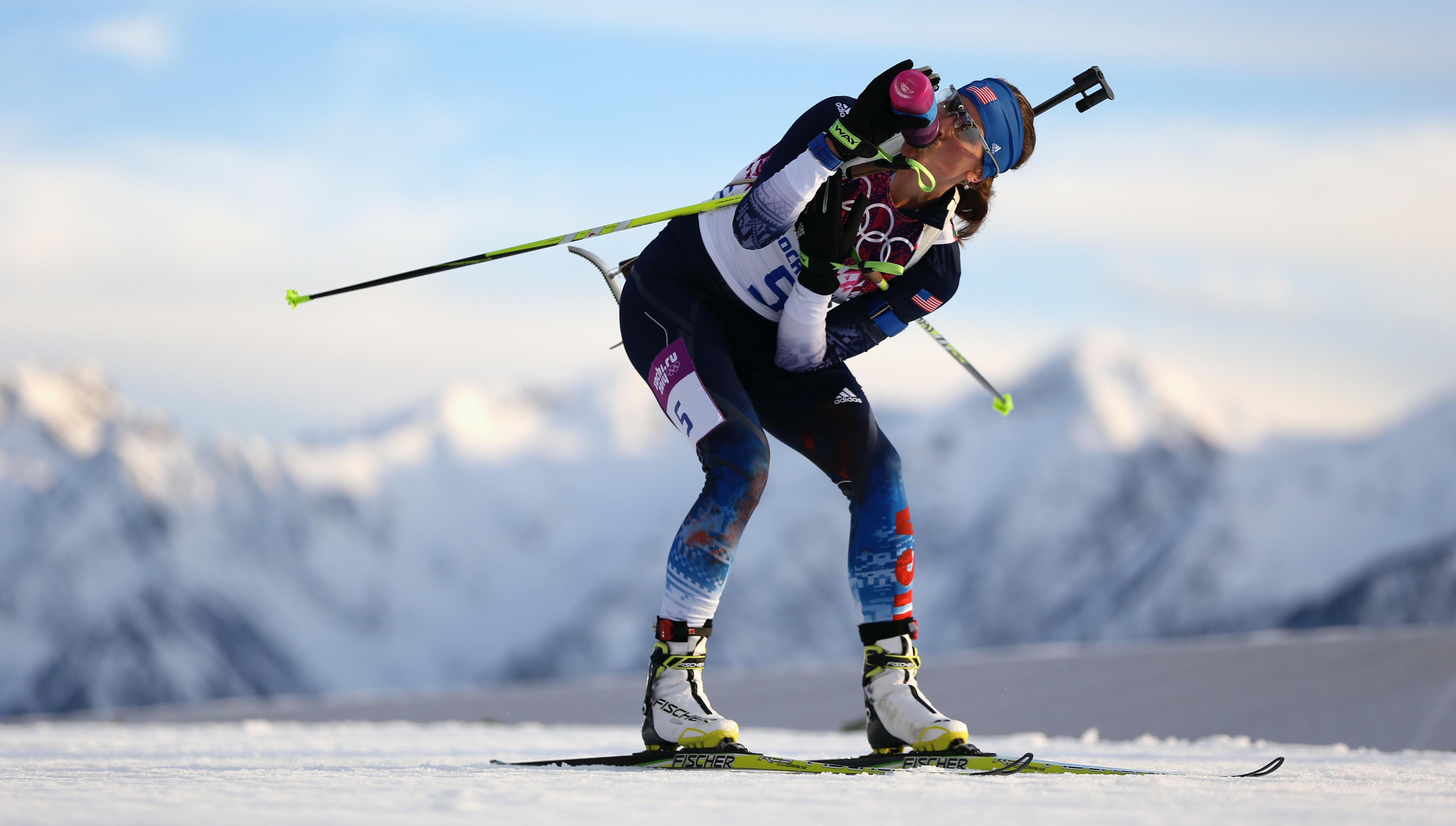 Biathlon athlete in a race on the snow, drinking from a bottle while skiing forward, poles on the side, mountains in the background.