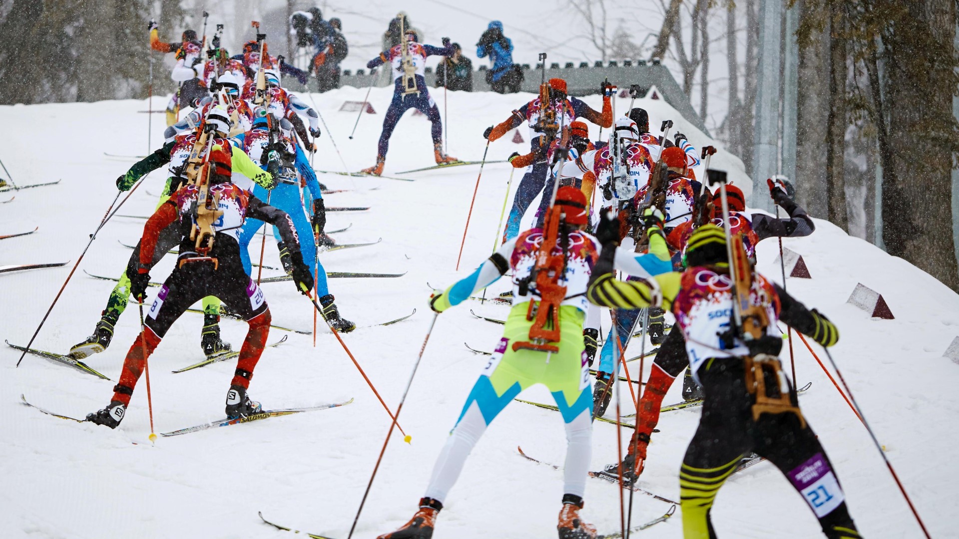 Several athletes seen from behind during an Olympic competition, rifle on the back, doing cross-country skiing.