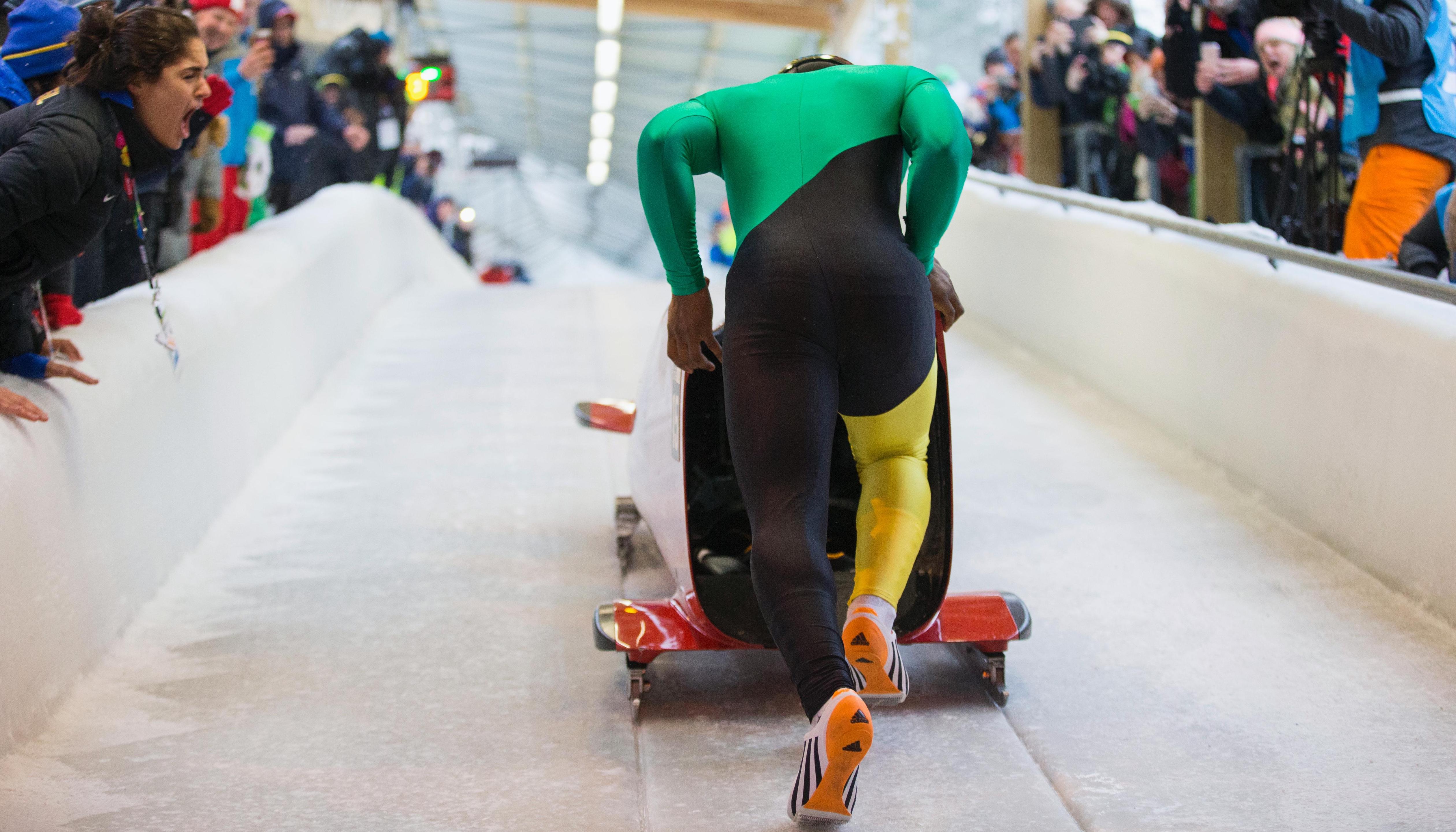 Athlete running on the ice-field while pushing the bobsleigh forward, seen from behind.