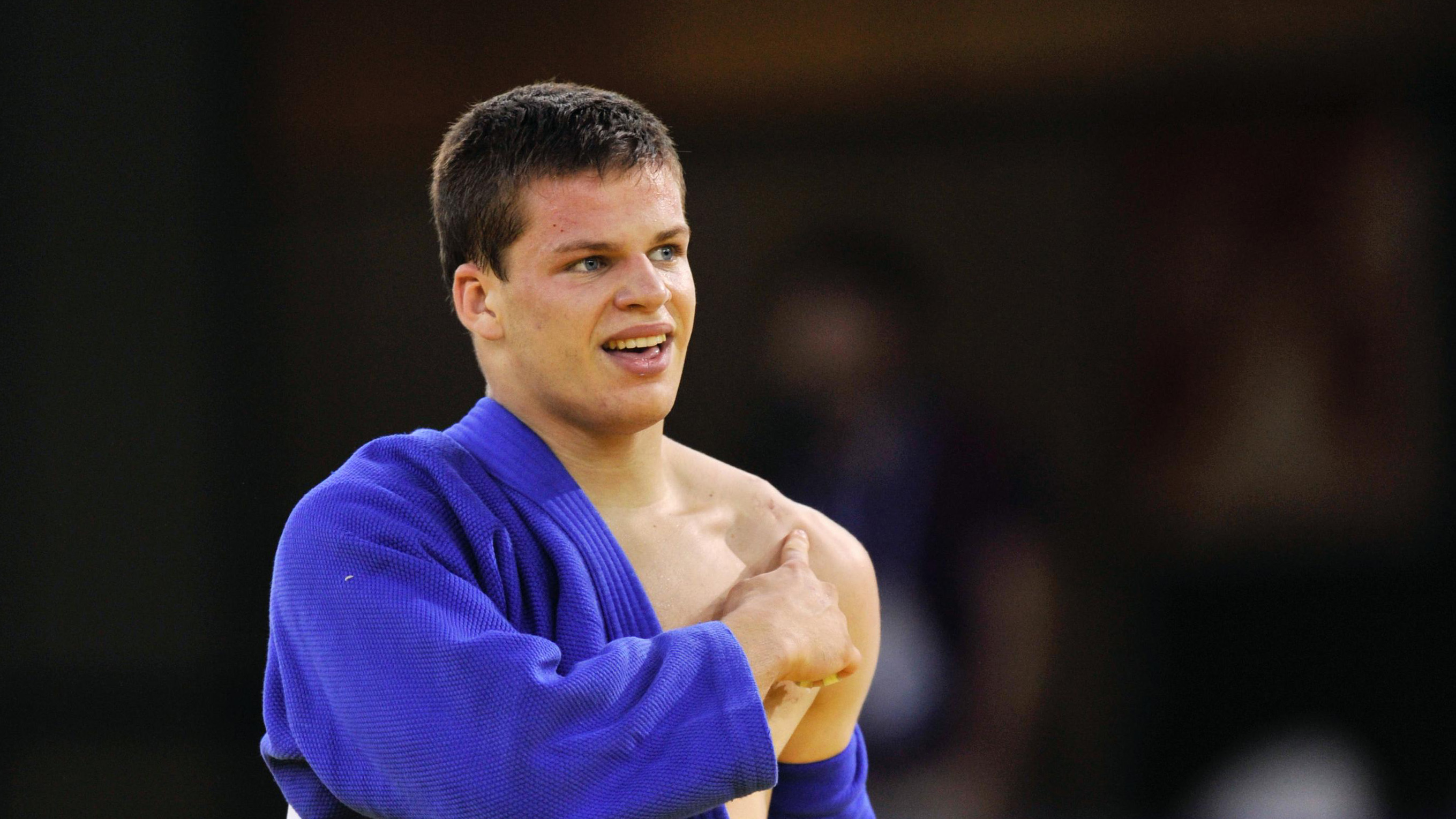 Athlete in a blue robe, pointing with his right hand on his left shoulder.