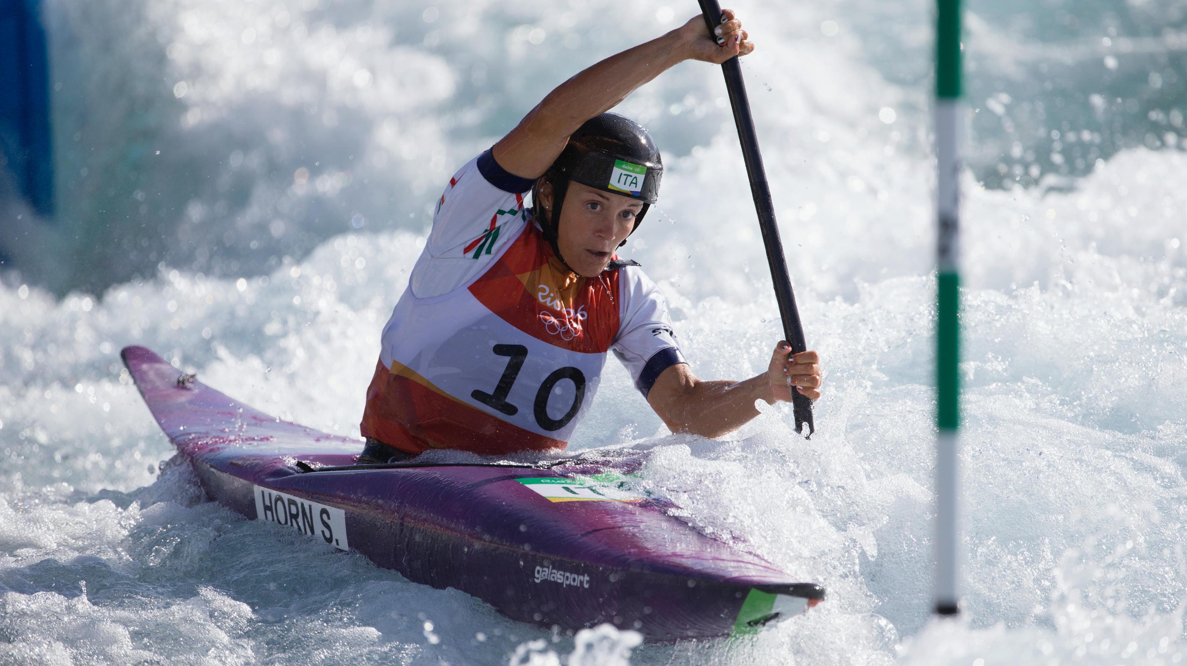 Athlete kayakking down a river, doing slalom between the poles, lots of water around.