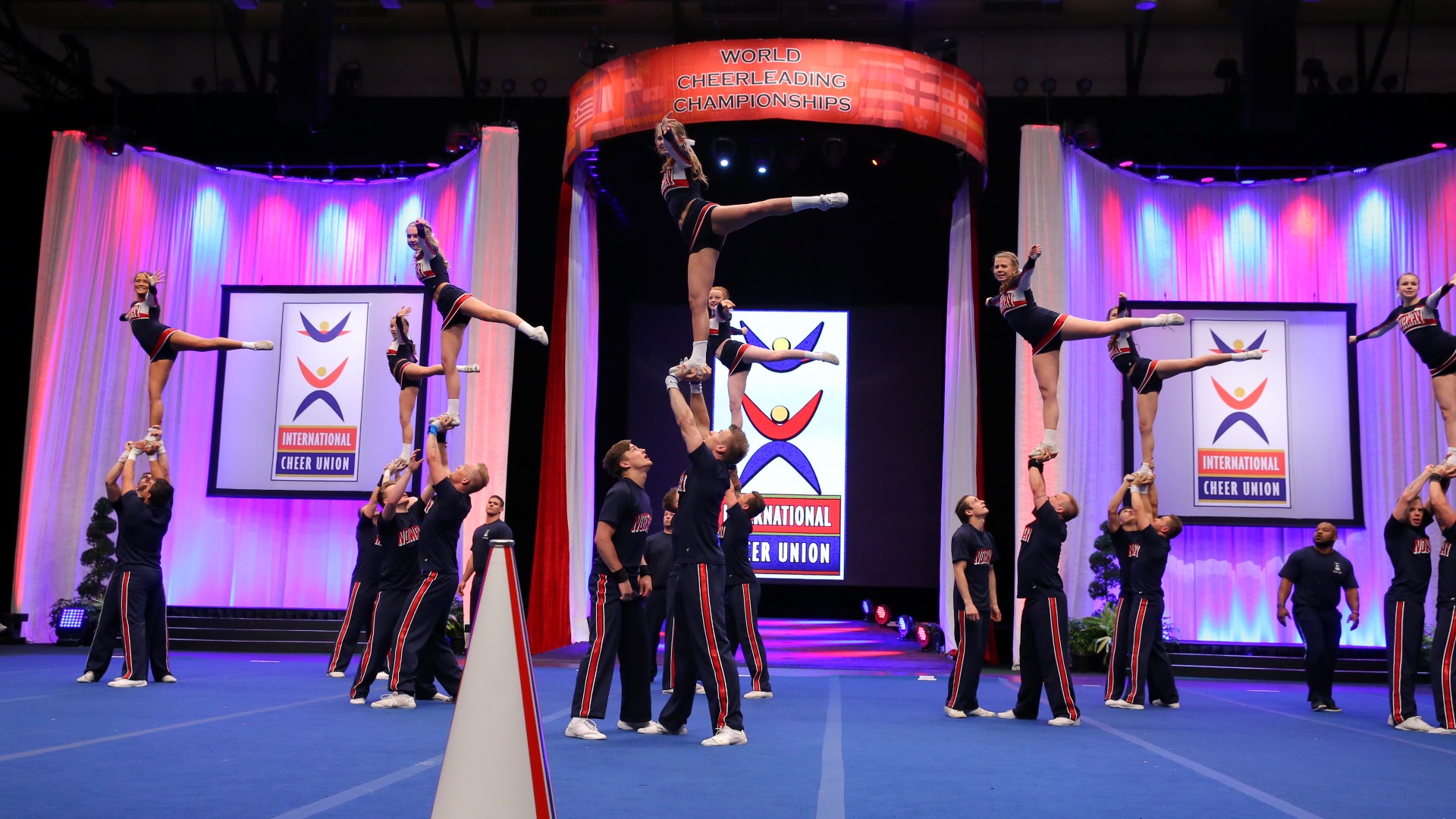 Several cheerleaders in a competition, that have lifted up many athlets that are standing on one leg while the other leg is standing straight out to the side.
