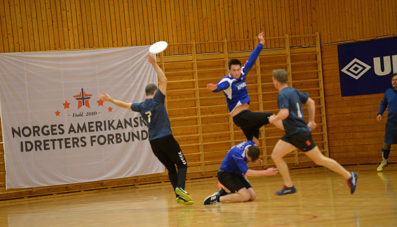 Several players in a disc-match indoors, one athlete have jumped in the air, disc flying through the room.