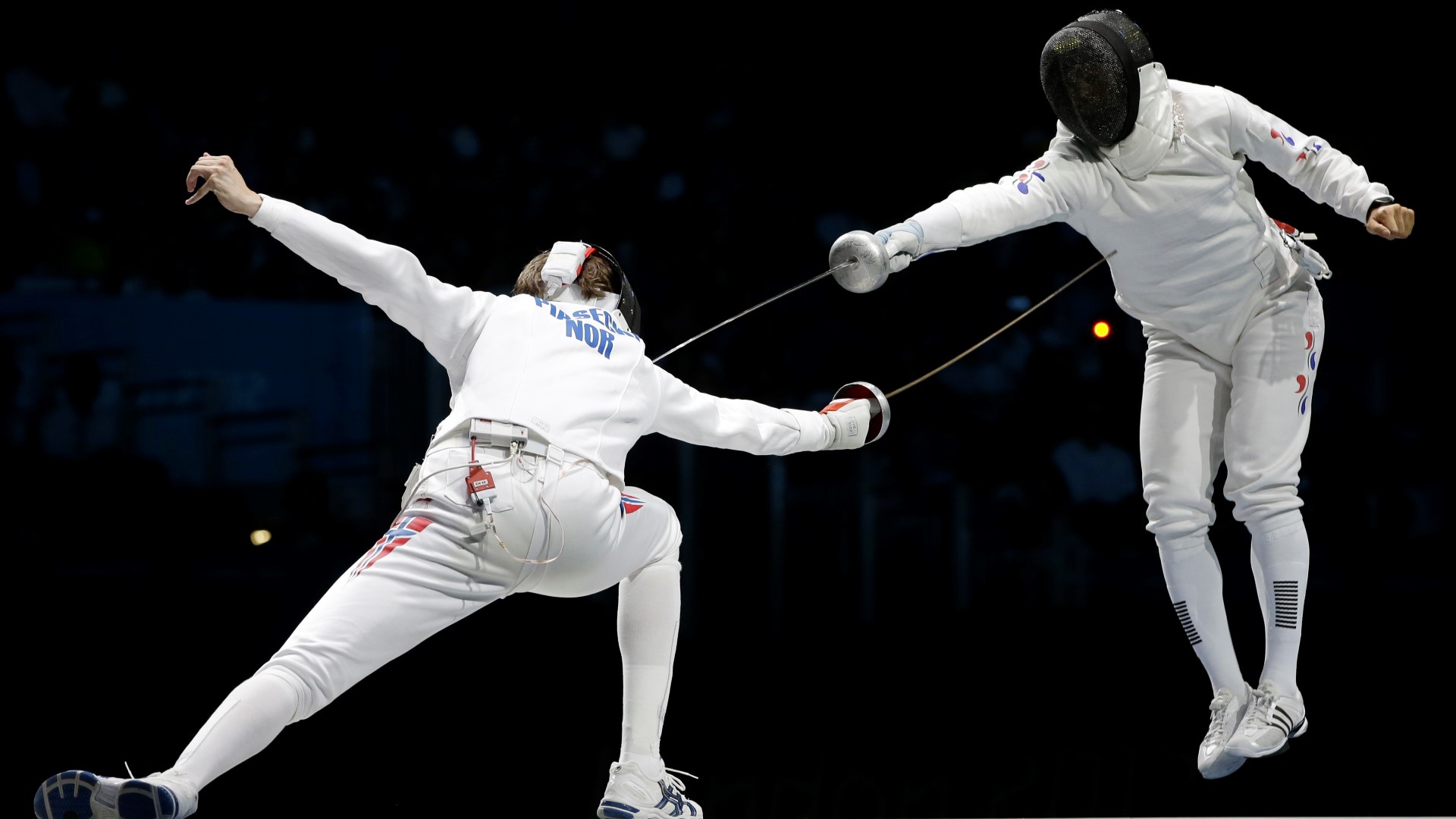 Two fencers in a match. One has jumped up and is in the air while he is pointing the cord on the other athlete. The other athlets is bendt forward, arm stretched out and the cord is pointing towards the athlete.