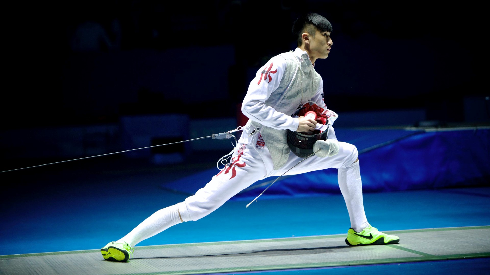 Fencer leaning forward, one leg behind the other leg bendt. Holding his cord with his left hand and the helmet in the right hand close to his body.