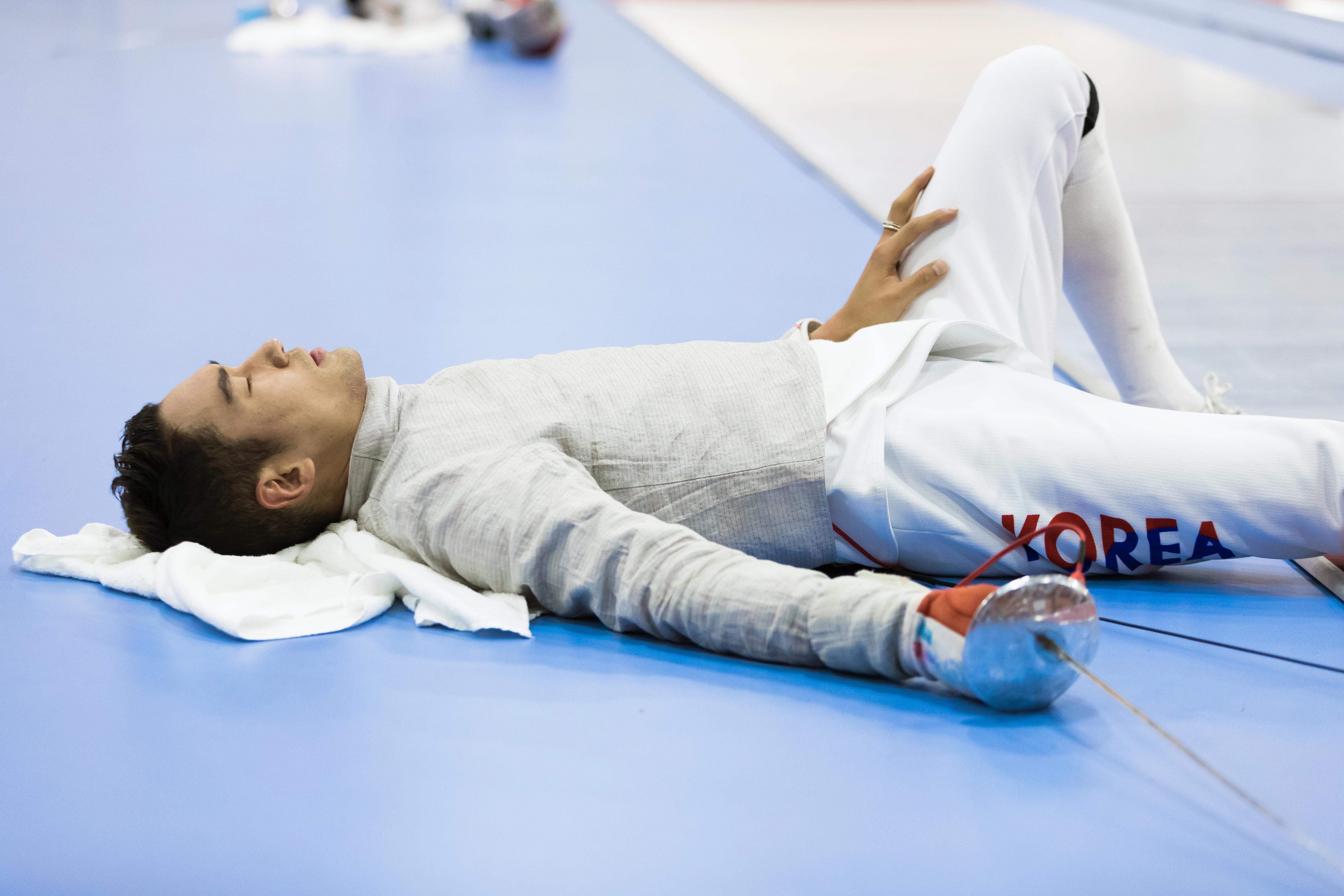 Fencer laying on the back on the ground, eyes cloesd and the cord in his right arm on the ground.