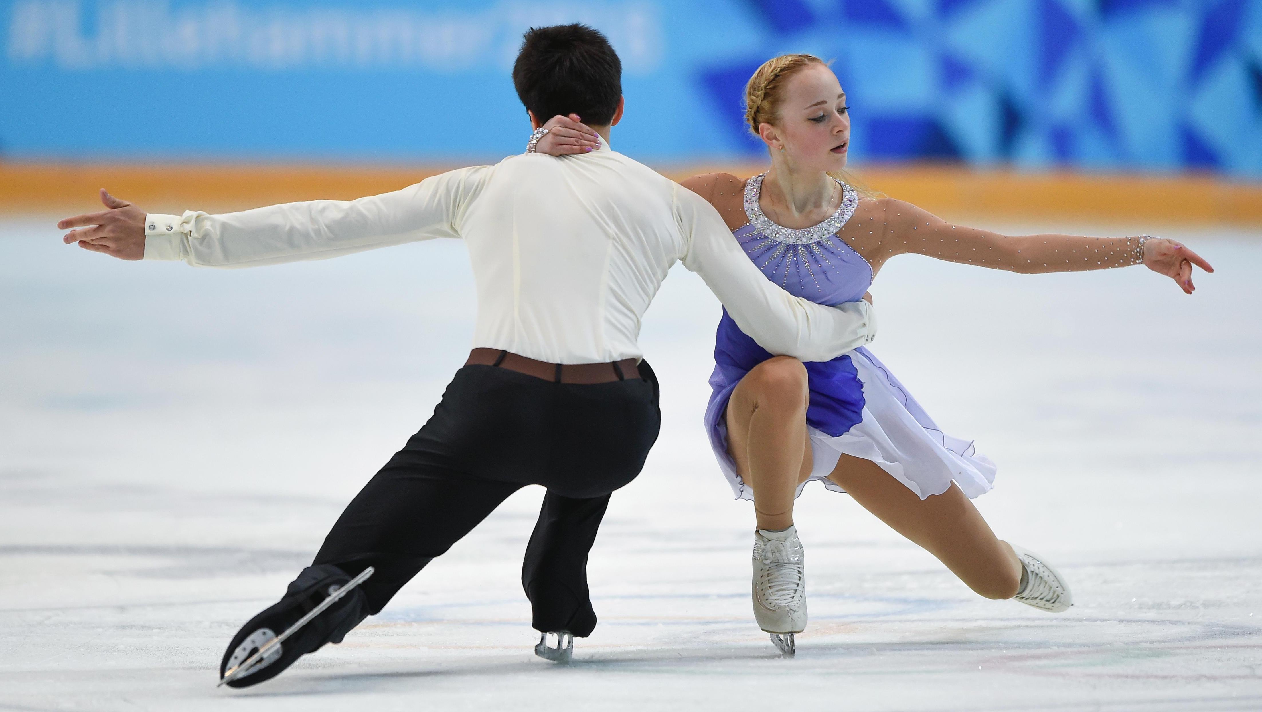 Two figure skaters on the ice in a competiton. One leg bendt and the other leg stretched out.