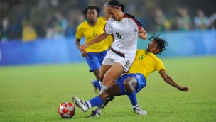 Footballers on the field, one athlete is tackling the other athlete who has the ball in front of her.