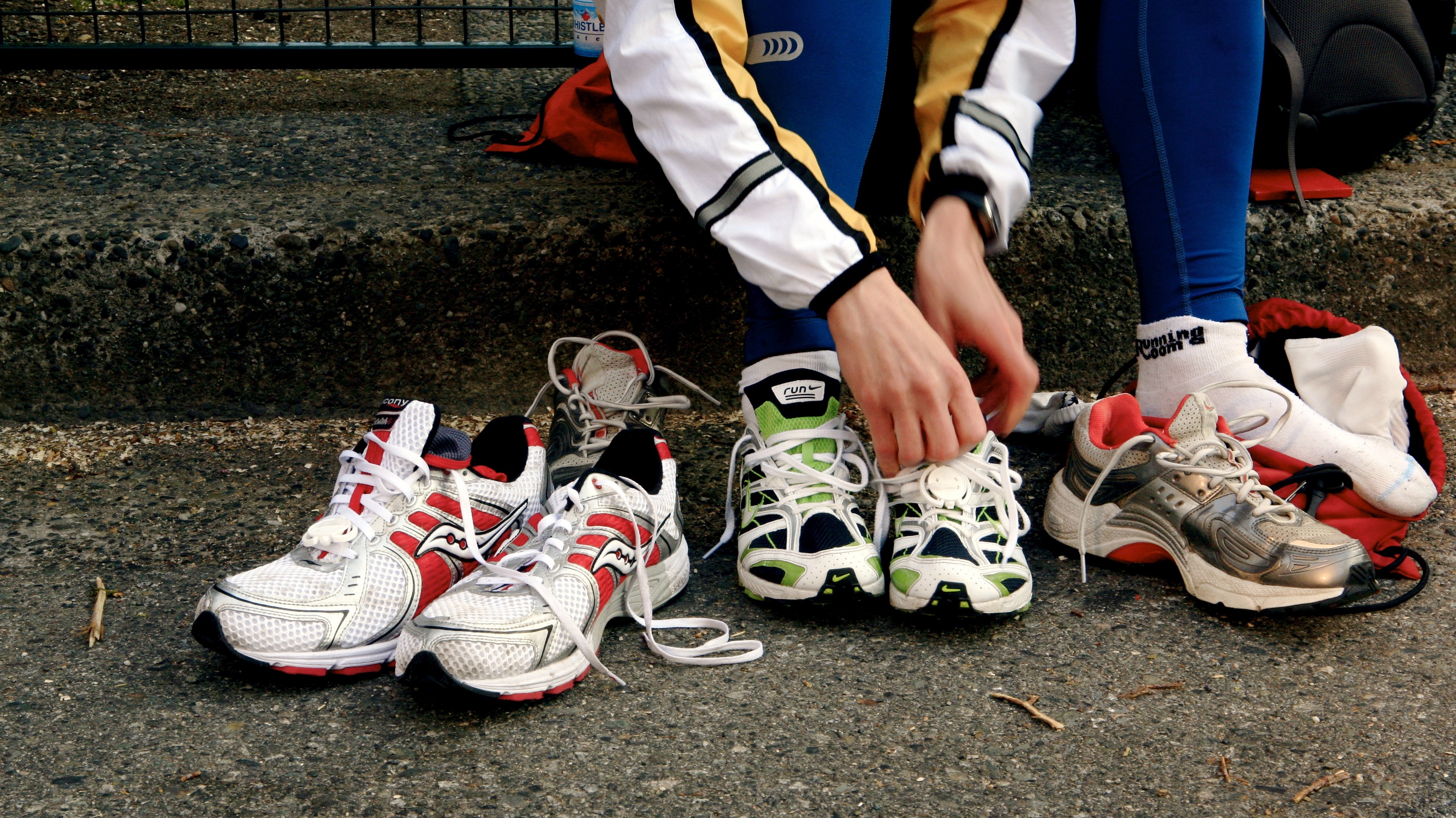 Several pairs of running shoes lined up, outside on the pavement, while an athlete is prepared to put one of the shoes on.
