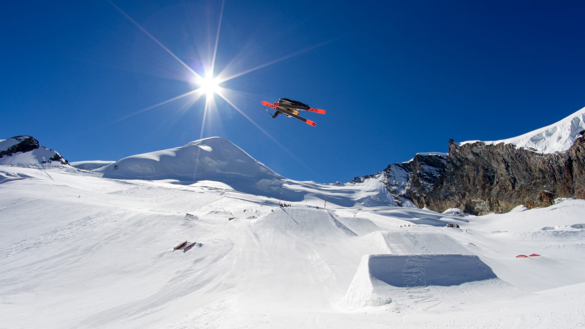 Freeskier high up in the air after a jump, sun and mountains in the background.