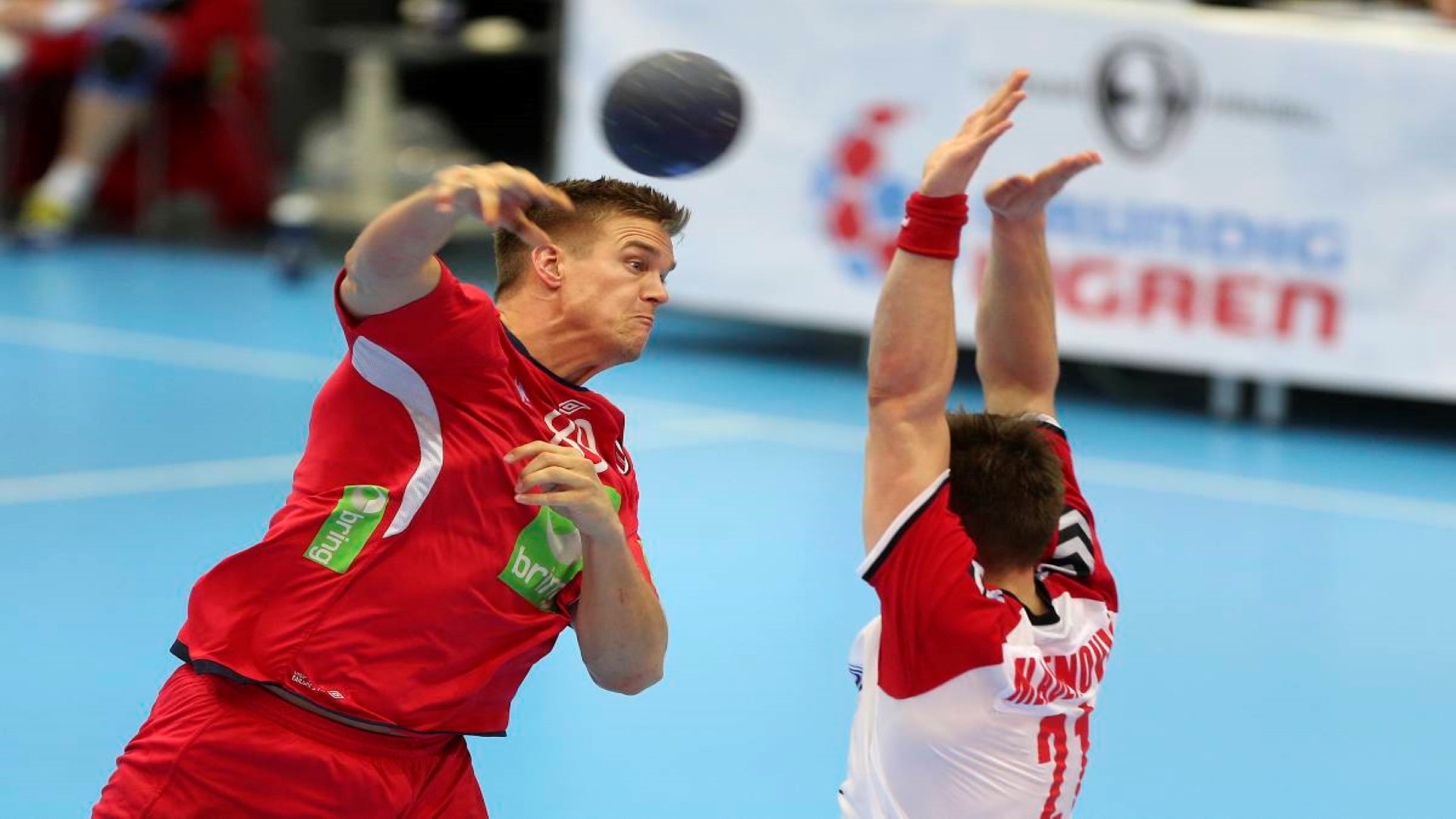 Two handballplayers in a match, one athlete has just thrown the ball while the other players is trying to block the shot with his arms up.