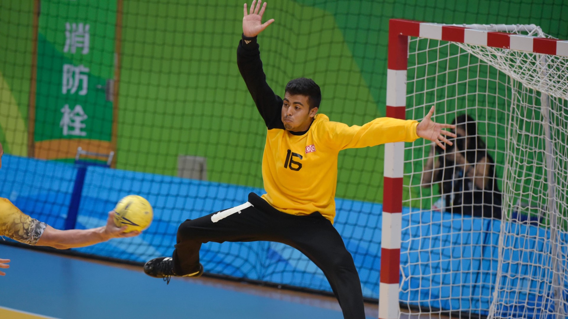 Handball goalkepper jumping up in the air in a star formation, while another athlete tries to shoot for the goal.