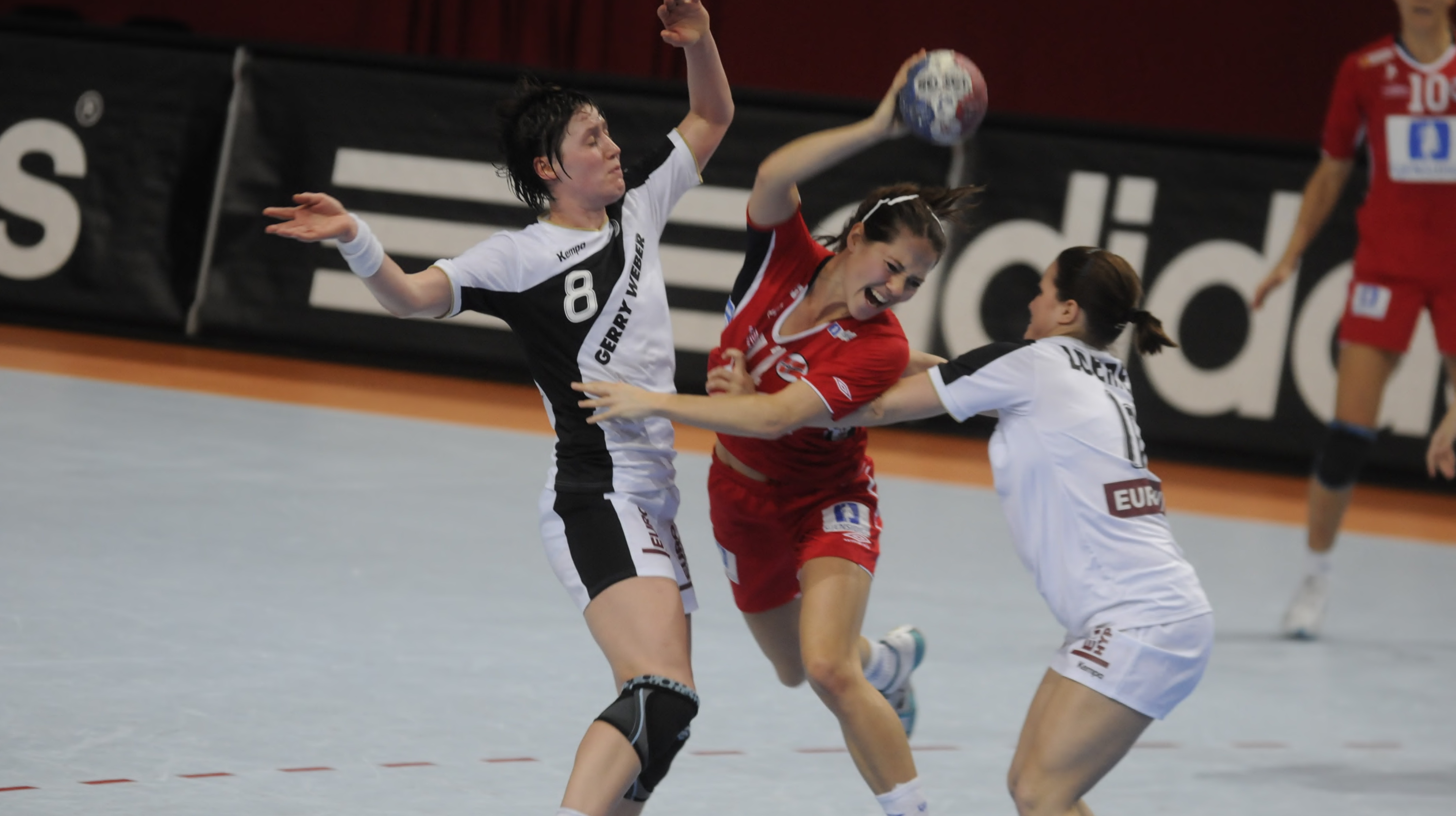 Three handballplayers in a match on the court, one of the athletes in the middle of two opponents trying to tackle her.