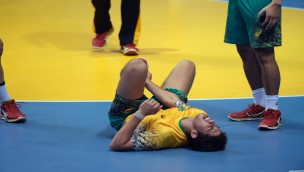 Athlete lying on the court in pain, holding his right hand on his leg. Other players around him.