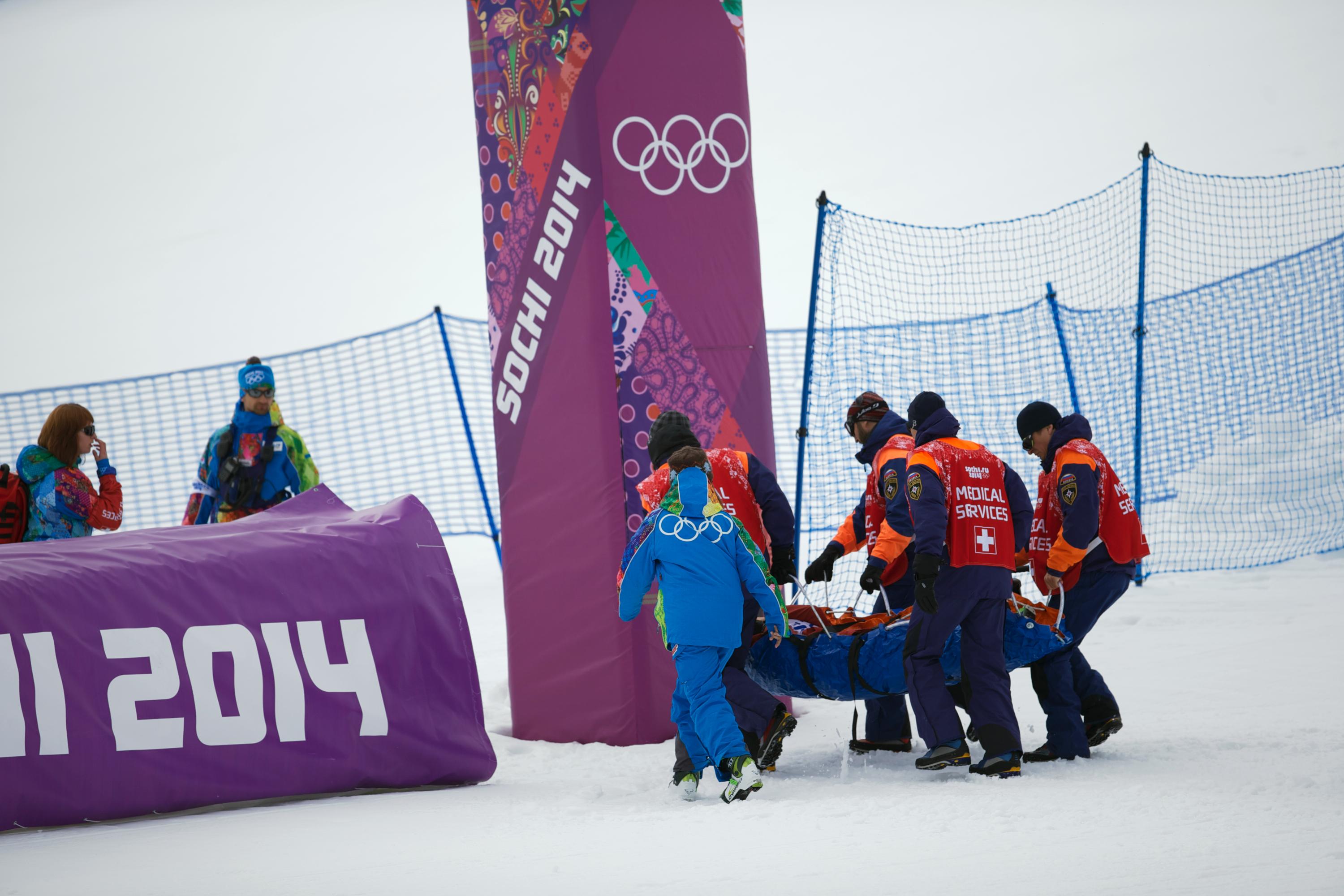 Several medical personel carrying an athlete of the competition field on a stretcher, snow all around. 