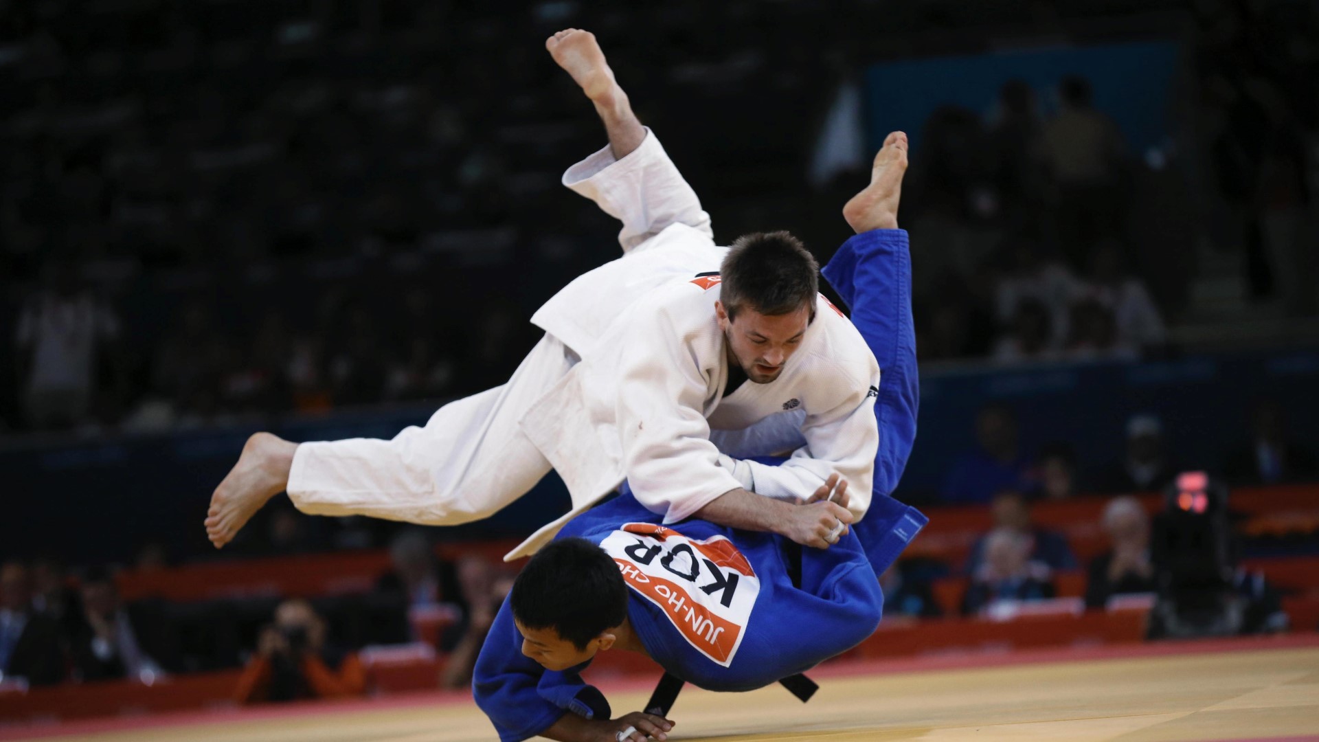 Two athlete in a Judo competition, one in the air about to jump on another athlete on the ground lying in his stomach.