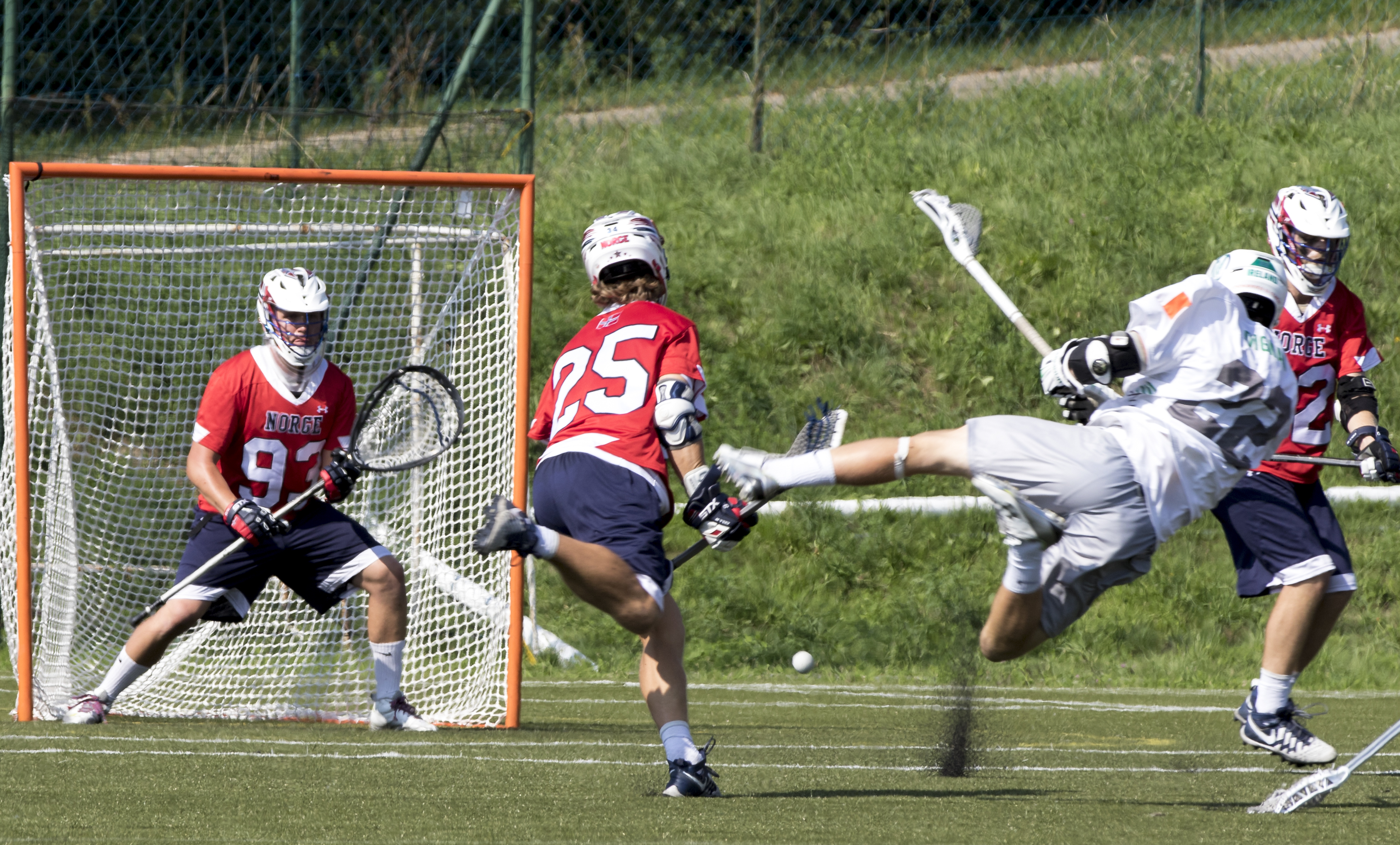 Lacrosse players in a match. One player lies in the air while making a shot towards goal. Goalkeeper looks forward on to the player in the air.