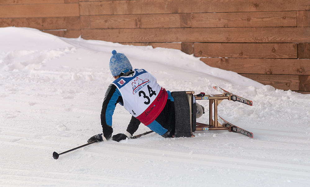 Sit-skier in a race, fallen over half-way on the side in the snow. Seen from behind.