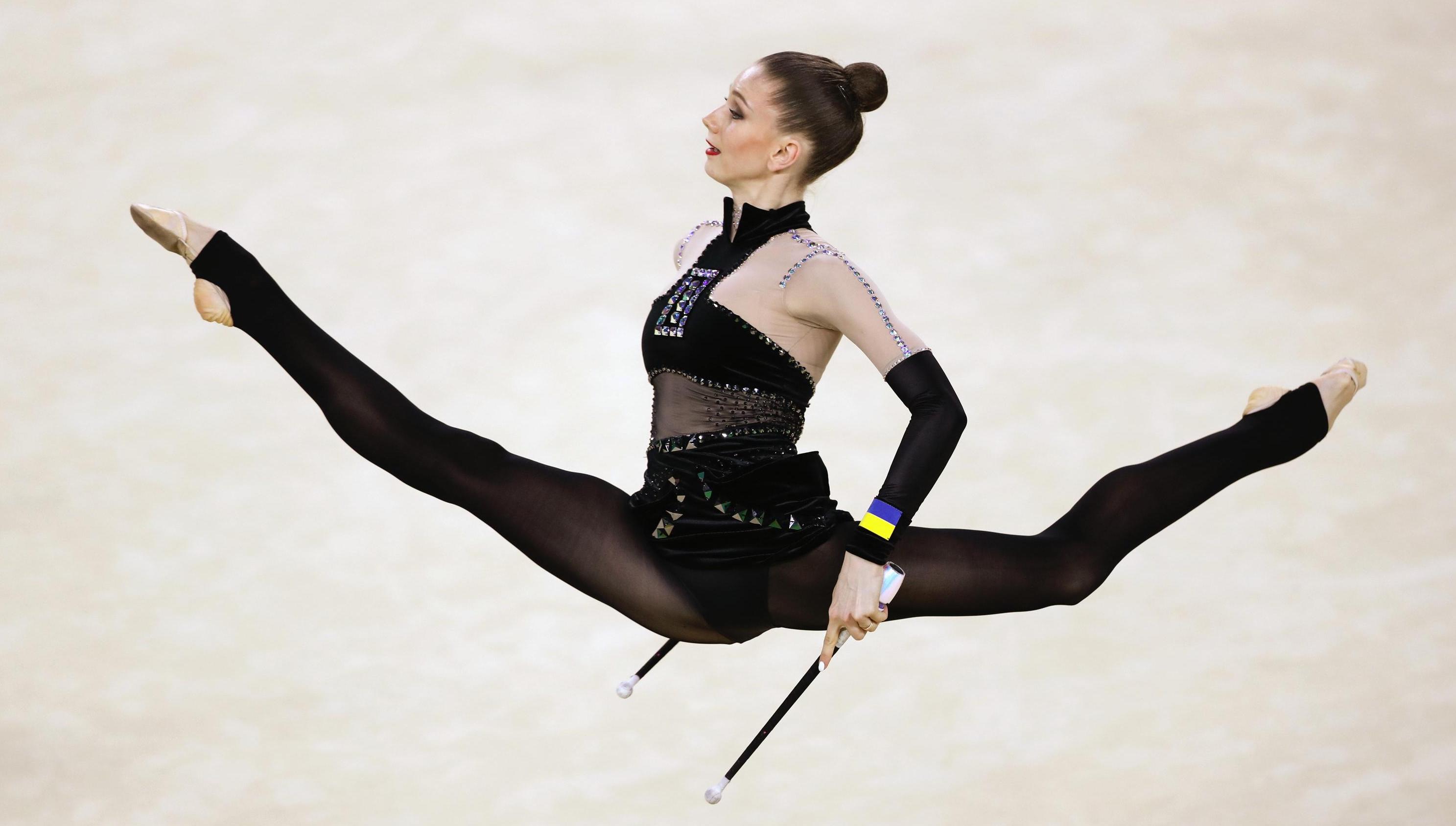 Athlete doing a split in the air, seen from the side. She is holing one stick in each hand.