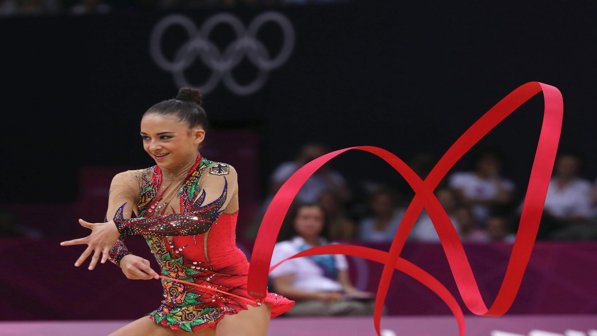 Athlete in a competition, holding a stick with a red long ribbon in her right hand. 