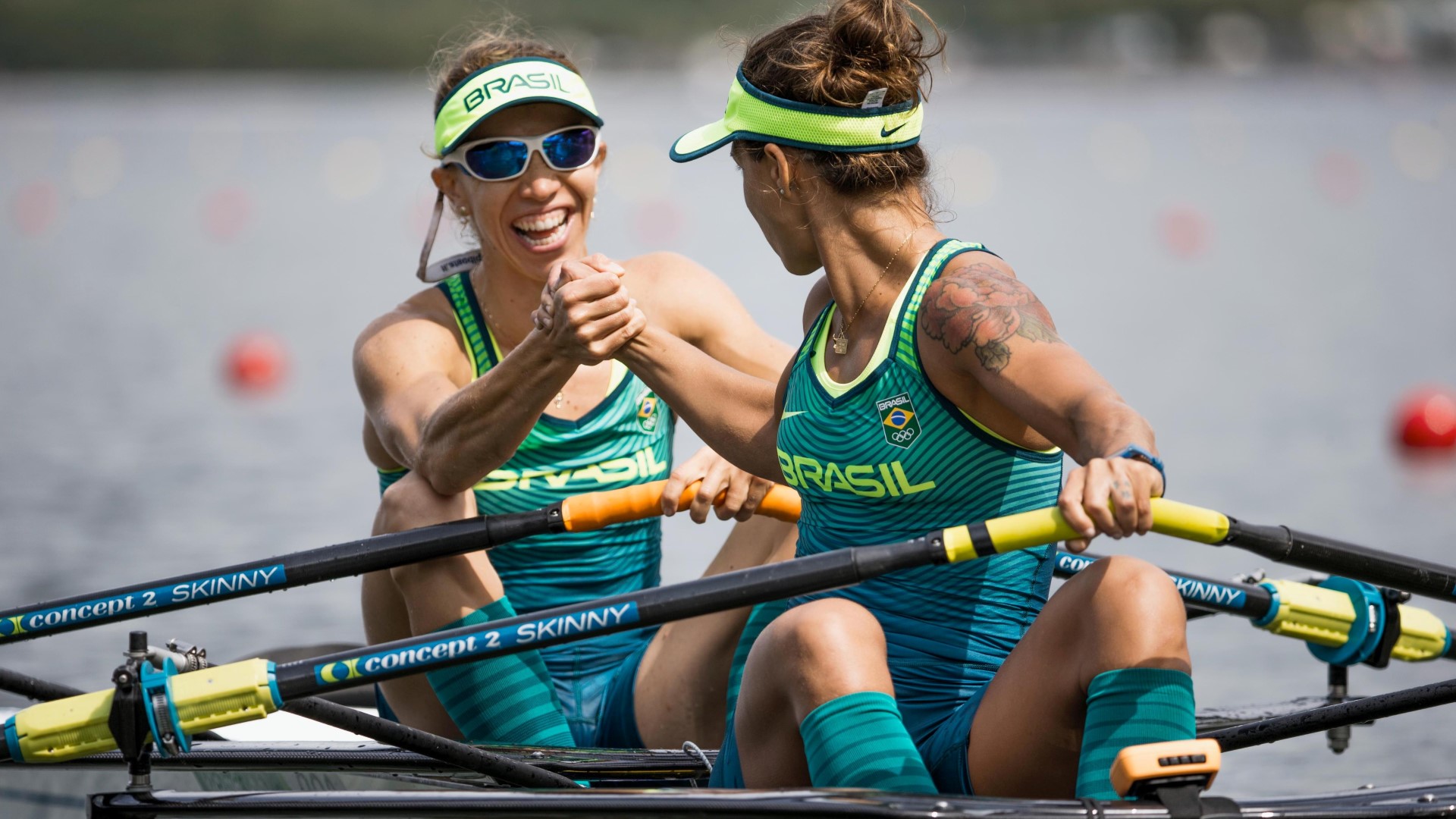 Two rowers in a boat, one turns back to the other and they are shaking hands and smiling.