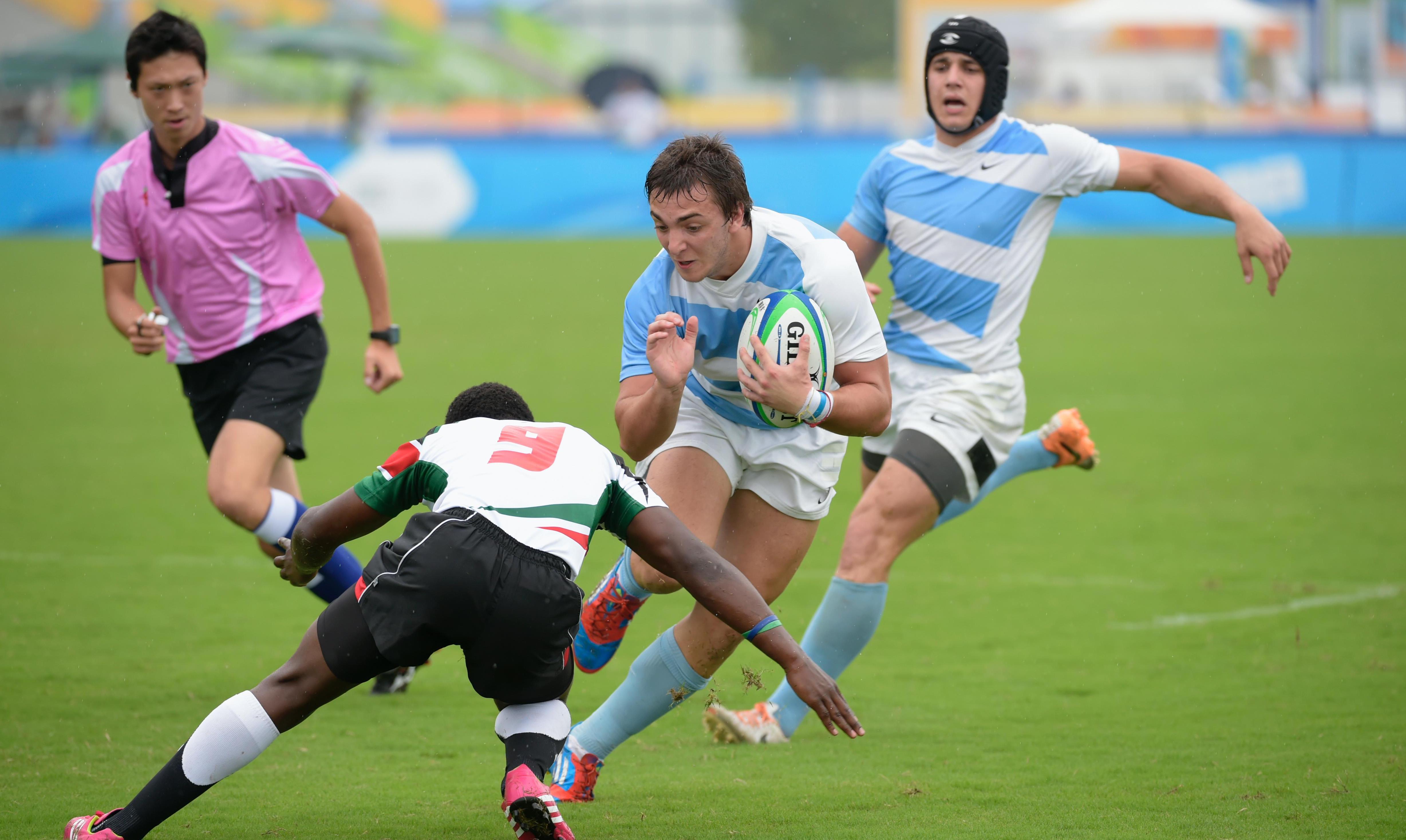 Three players and a referee in a rugby match. One of the players has the ball and an opponent is trying to tackle him.