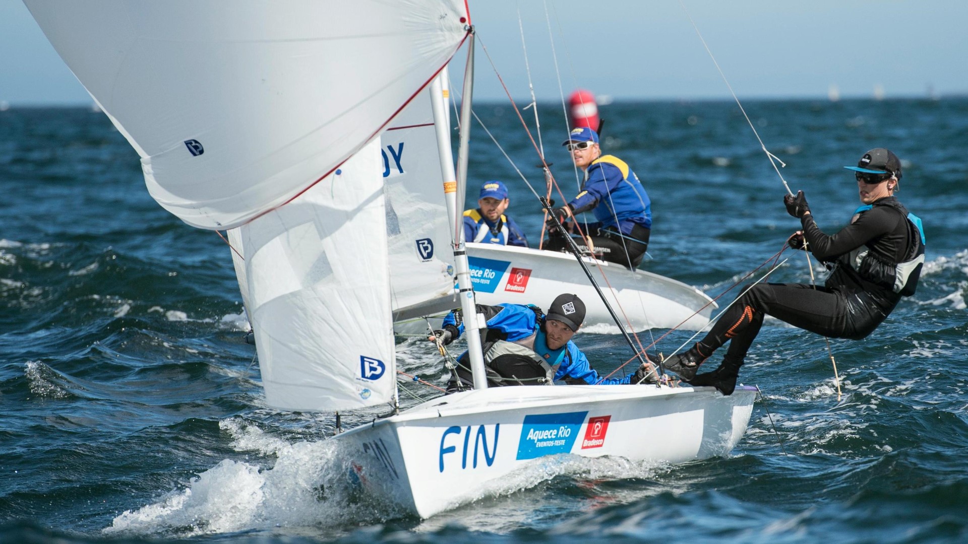 Two sailboats with two athletes in each boar, one in front of the other. One athlete hanging on the outside of the boar, holding on to the ropes. 
