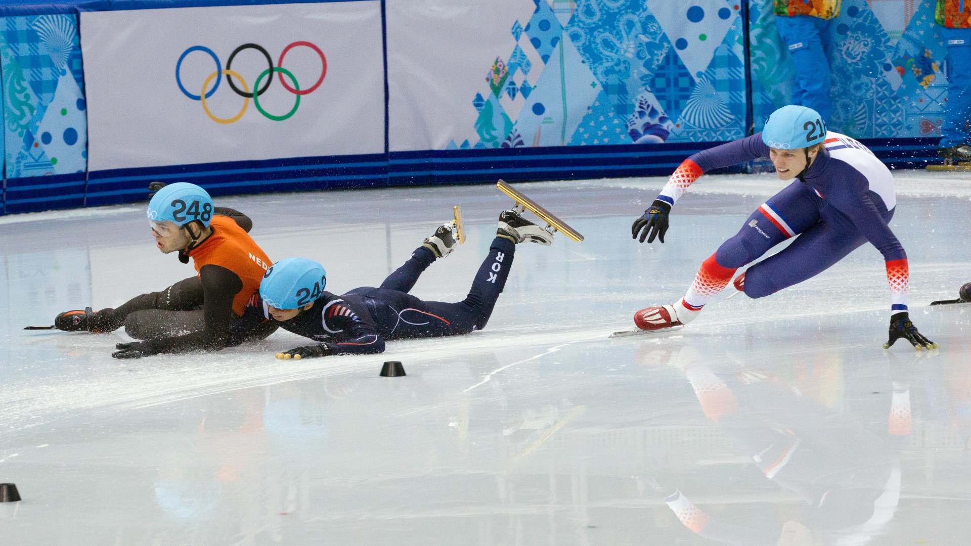 Three athletes in short track speed skating in a turn. Two have fallen down on the ice.
