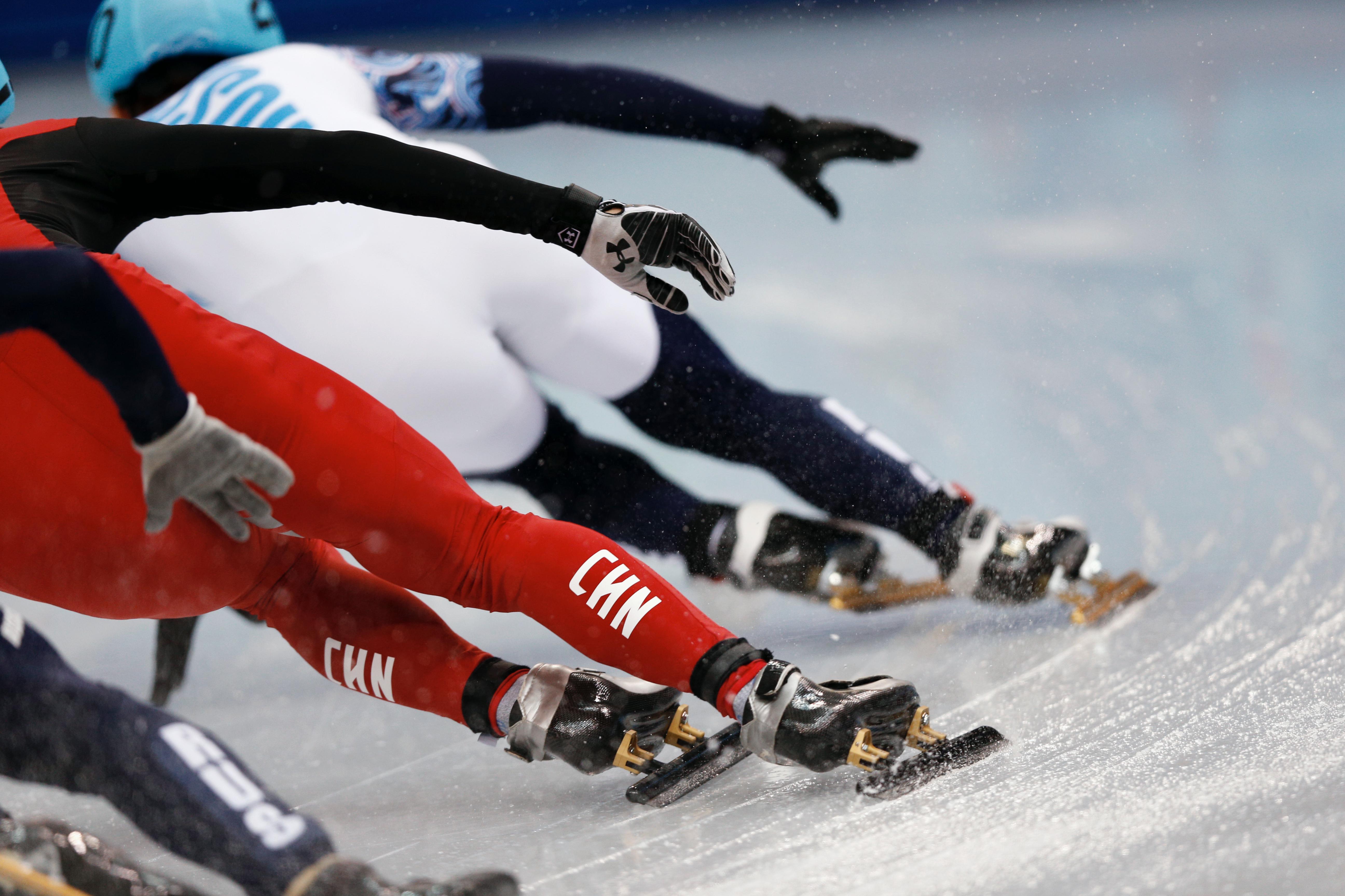Short track speed skaters seen from behind in a turn. See the skates on the ice, and their hand out to the side.