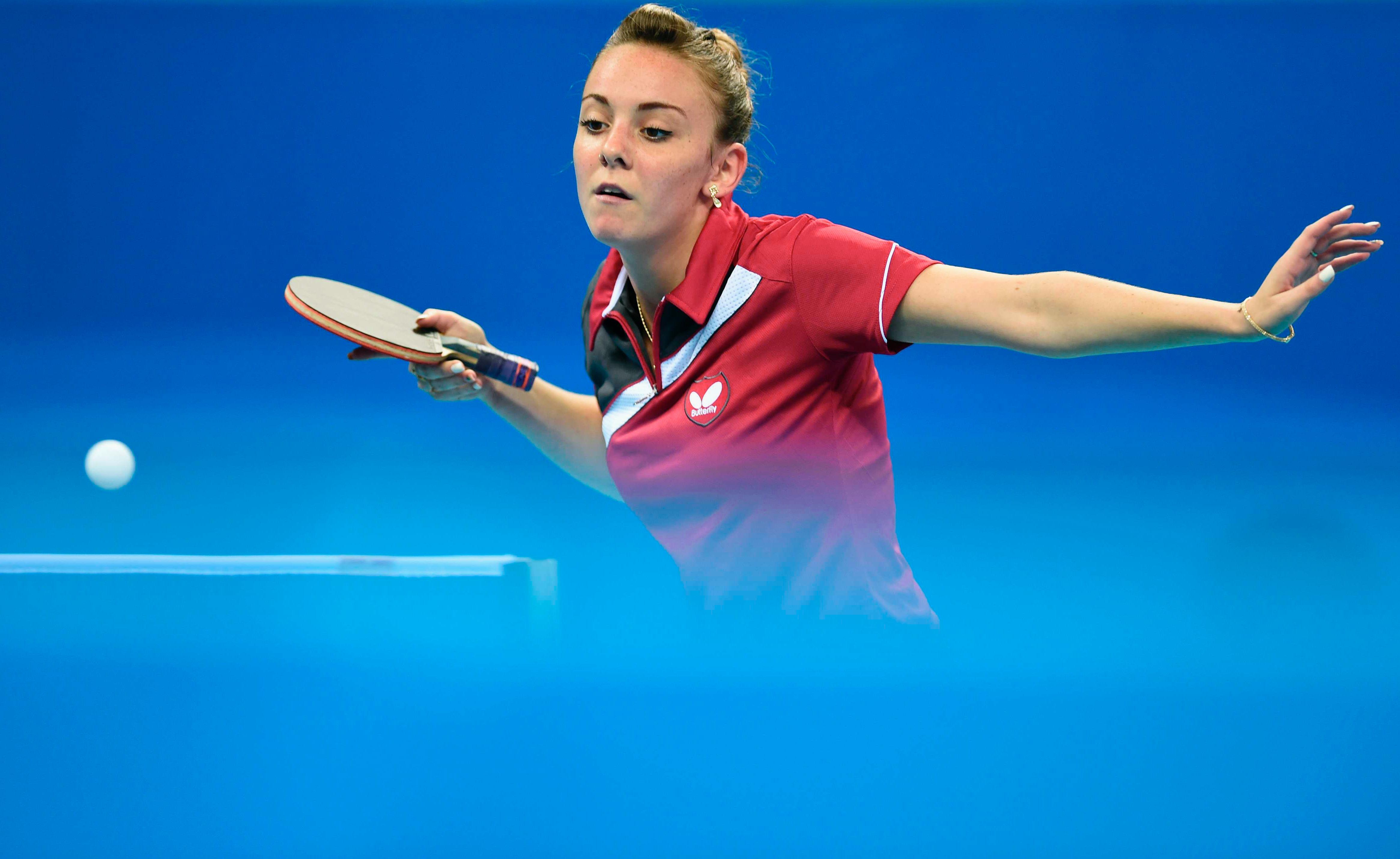 Blue background and some in the front. Athlete with the racket in her right hand, looking at the ball that is in the air.