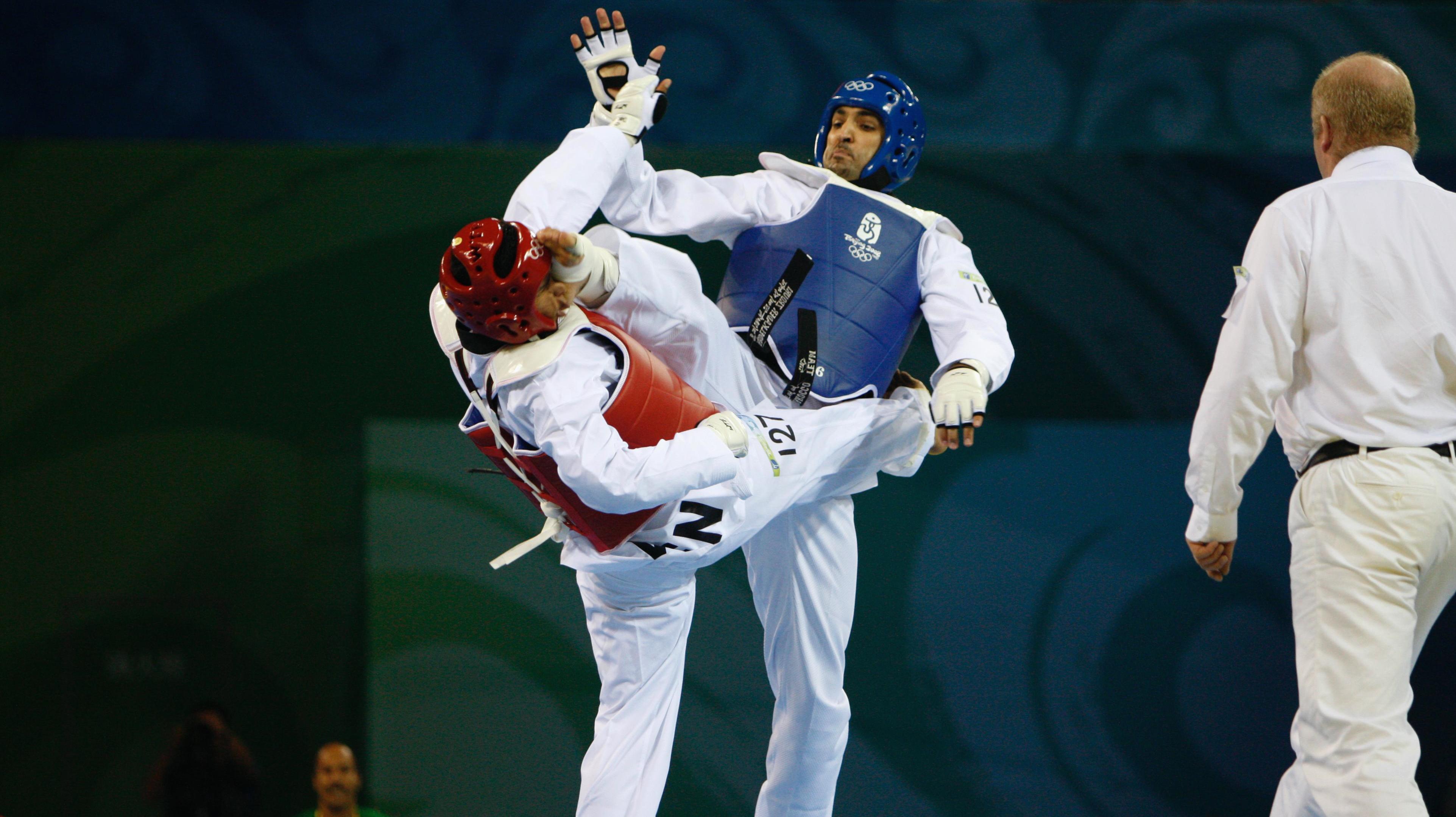 Two taekwondo athletes in a match, both kicking each other at the same time. Referee standing next to them observing.