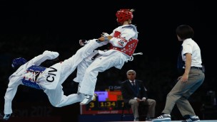 Two athletes in a teakwondo match, one athlete kicking the other athlete while they are both in the air. Referee is standing on the right watching them.