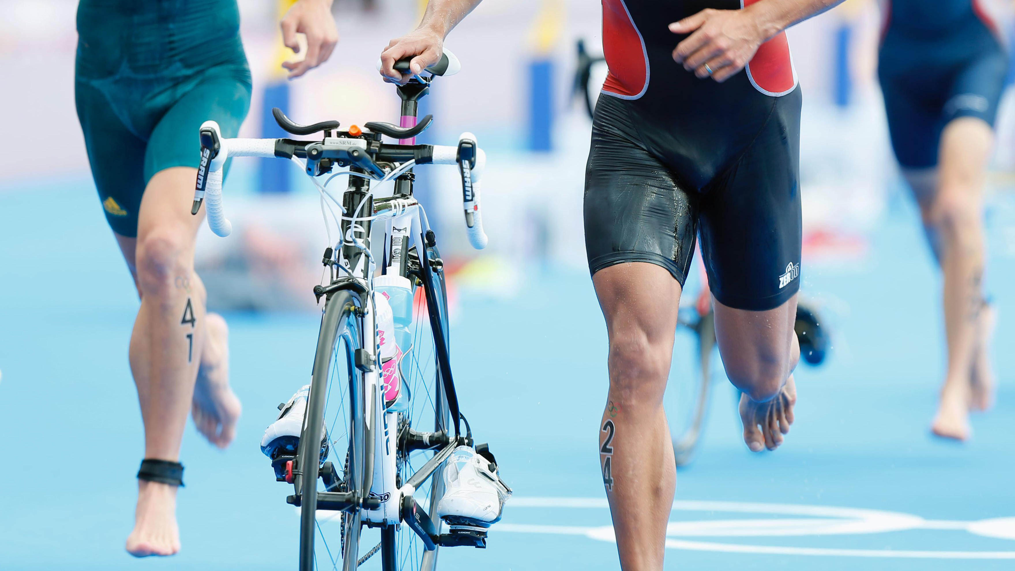 Close up of two triathletes, only seing the thigh, legs and feet. One of them is running with his bike, holding it with his right hand.