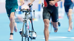 Close up of two triathletes, only seing the thigh, legs and feet. One of them is running with his bike, holding it with his right hand.