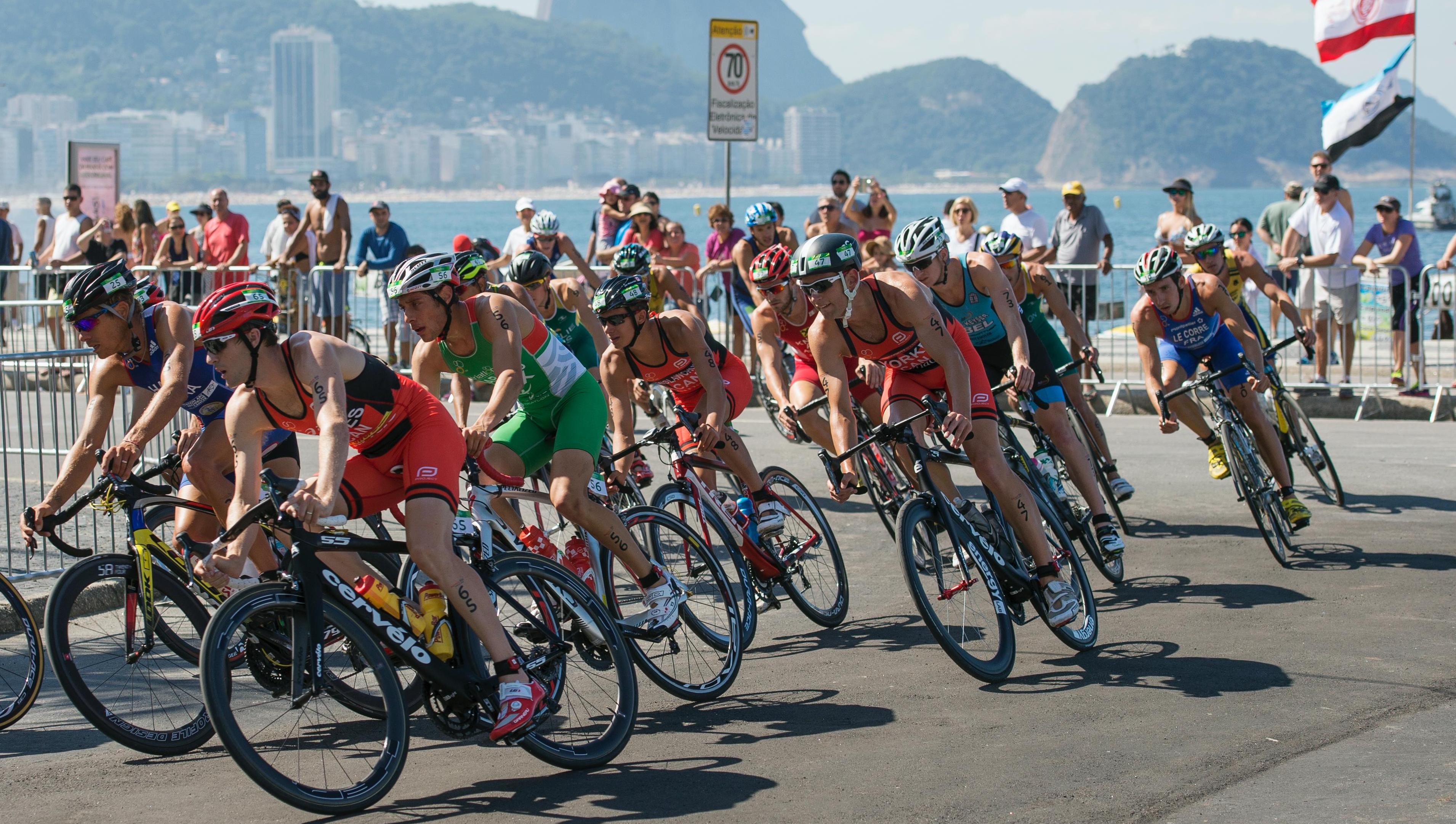 Several cyclists in an Olympic competition, in a turn, sea and mountain in the background.