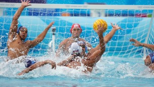 Several players in the pool during a water polo match. One athlete has the ball in his right hand and making a shot towards the goal.