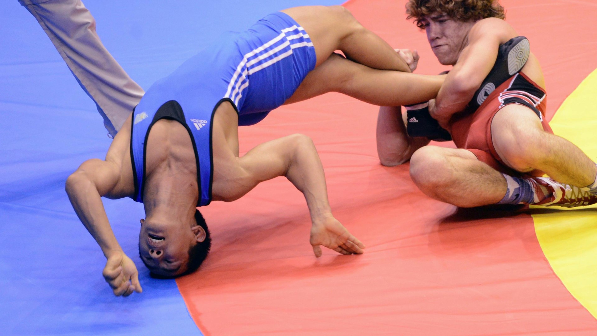 Two wrestlers in a match on the court. One is holding the other wrestlers foot, while the other athlete are falling down and backwards with the head on the court.