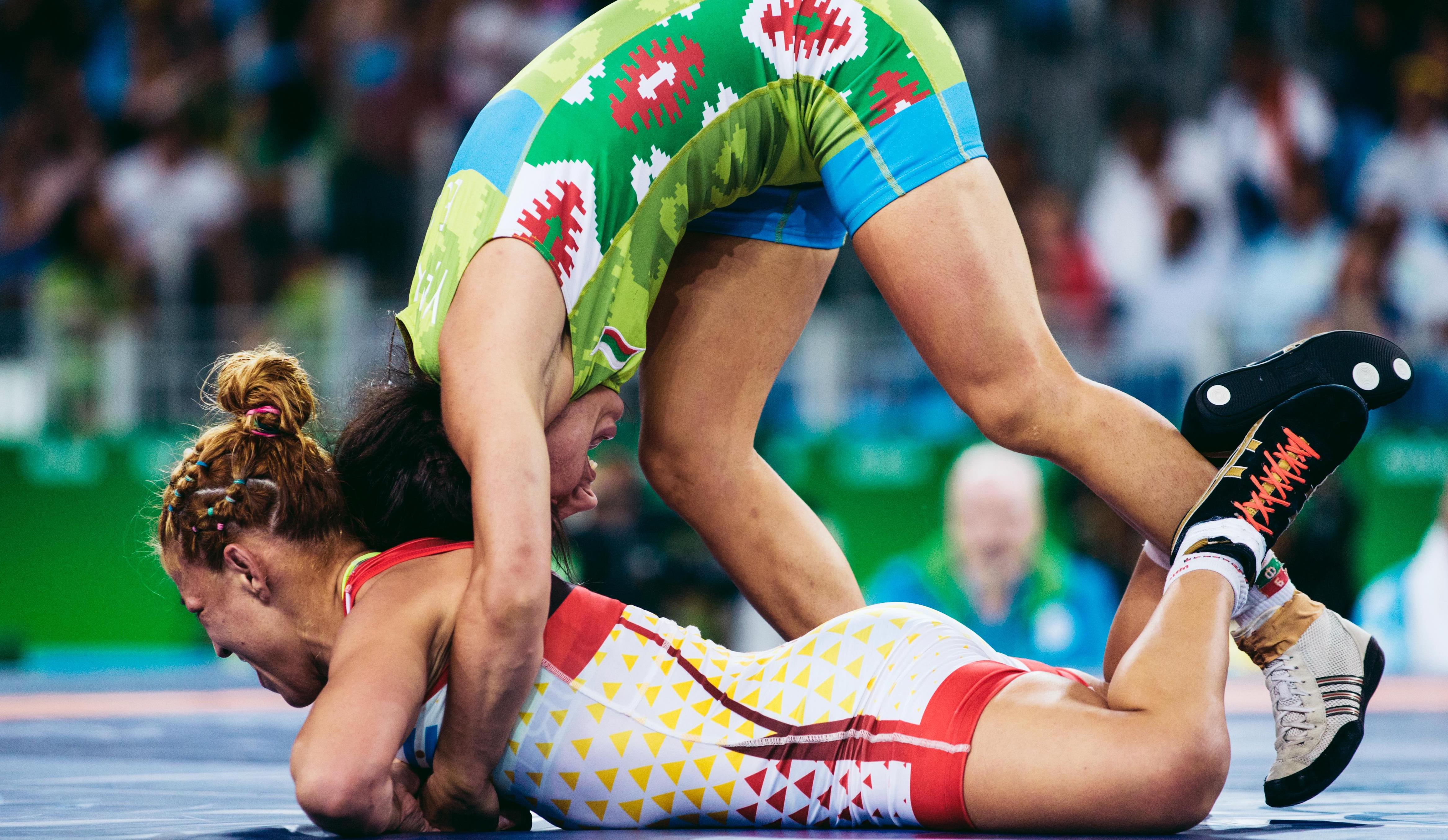 Two wrestlers in a match, one athlete lying on the stomach while the other athlete is standing behind with her arms around the other athlete.